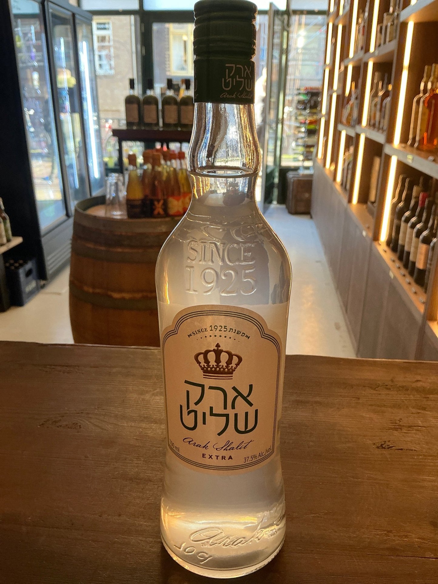 Clear glass bottle of Arak Shalit Extra premium artisan Israeli arak with embossed crown logo and black cap on wooden.