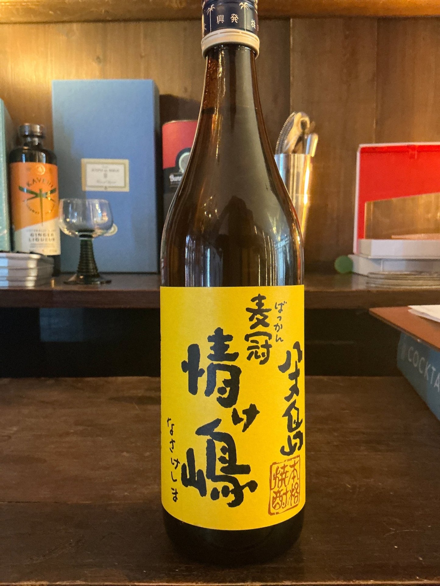 Bakkan Nasake Shima Shochu brown glass bottle with bright yellow label featuring Japanese calligraphy, artisan small batch.