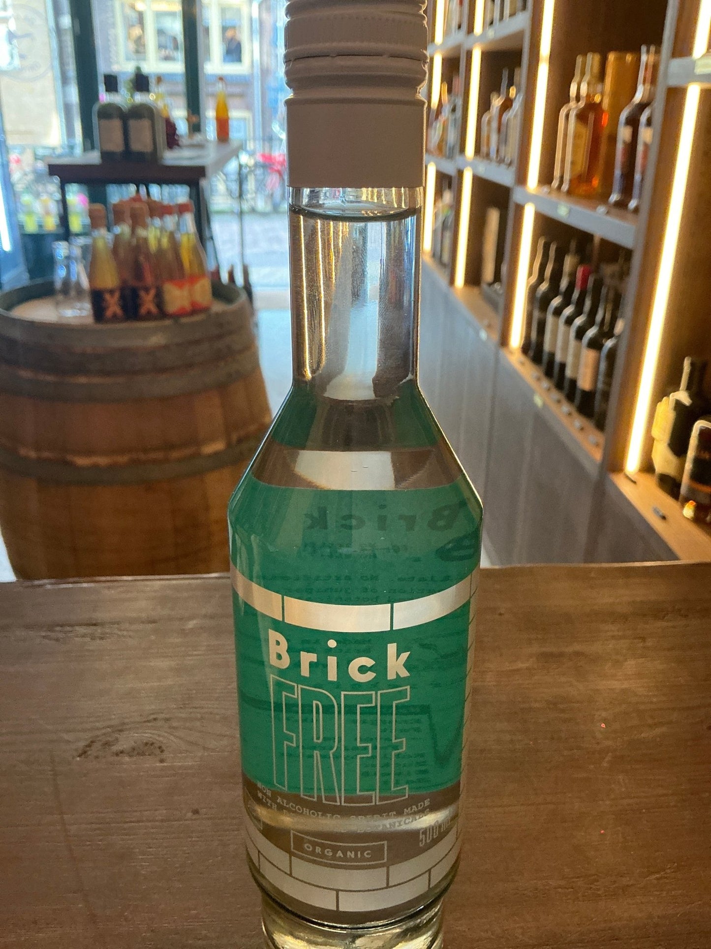 Clear glass bottle of Brick Free non-alcoholic gin with teal and white botanical-themed label, displayed on wooden counter.