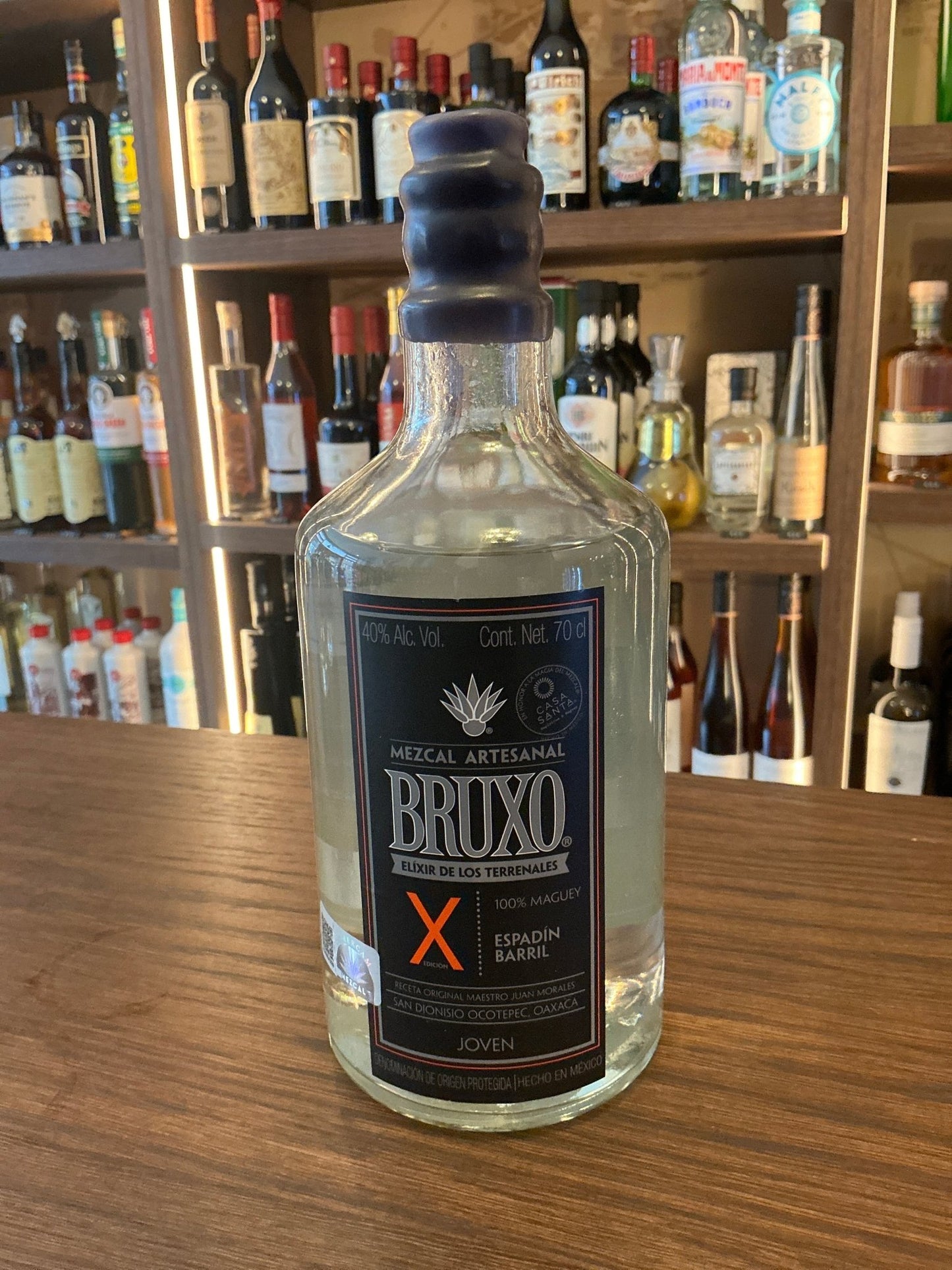 Bruxo X Espadin Mexican mezcal bottle with dark label and black wax-sealed cap on wooden bar, premium small batch spirit.