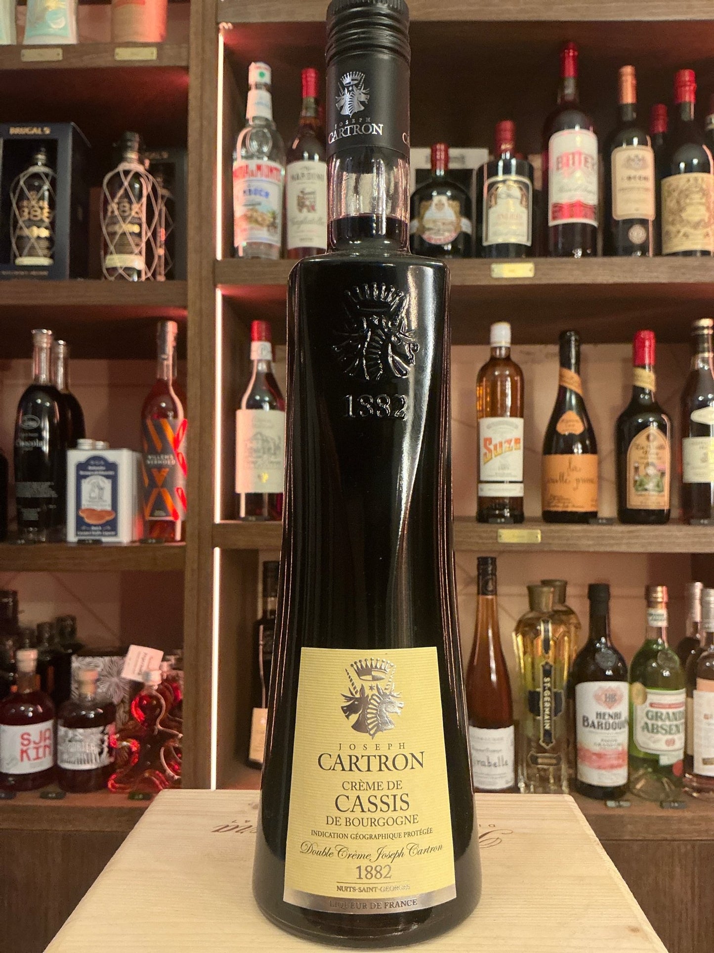 Tall dark glass bottle of Carton Crème de Cassis liqueur with botanical and artisan styling on label, displayed against.