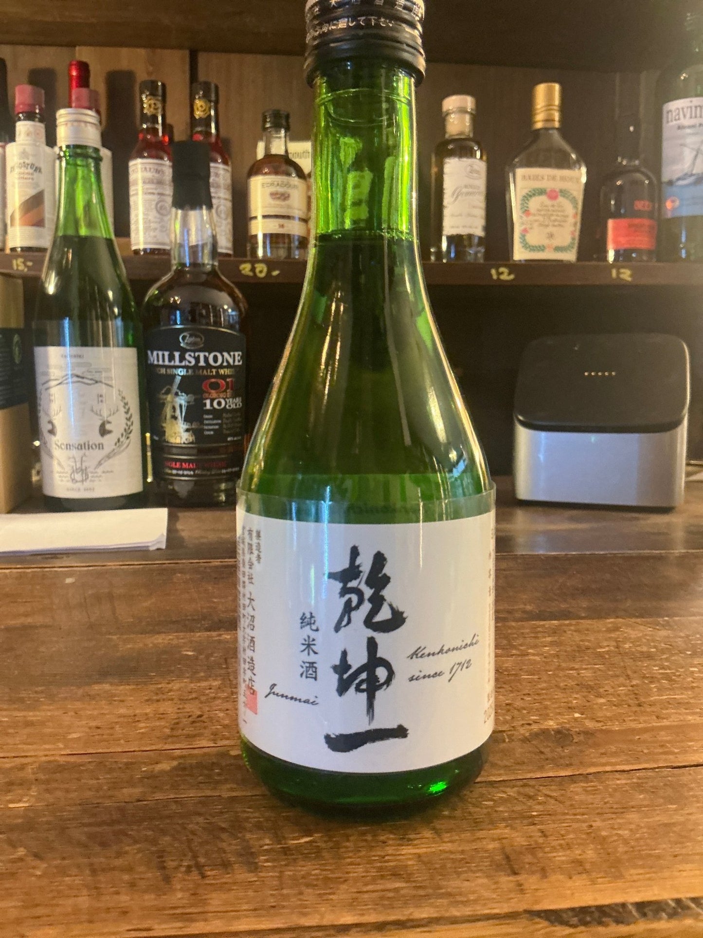 Green glass bottle of Kenkonichi Junmai-shu sake with white label featuring Japanese calligraphy on wooden bar counter,.