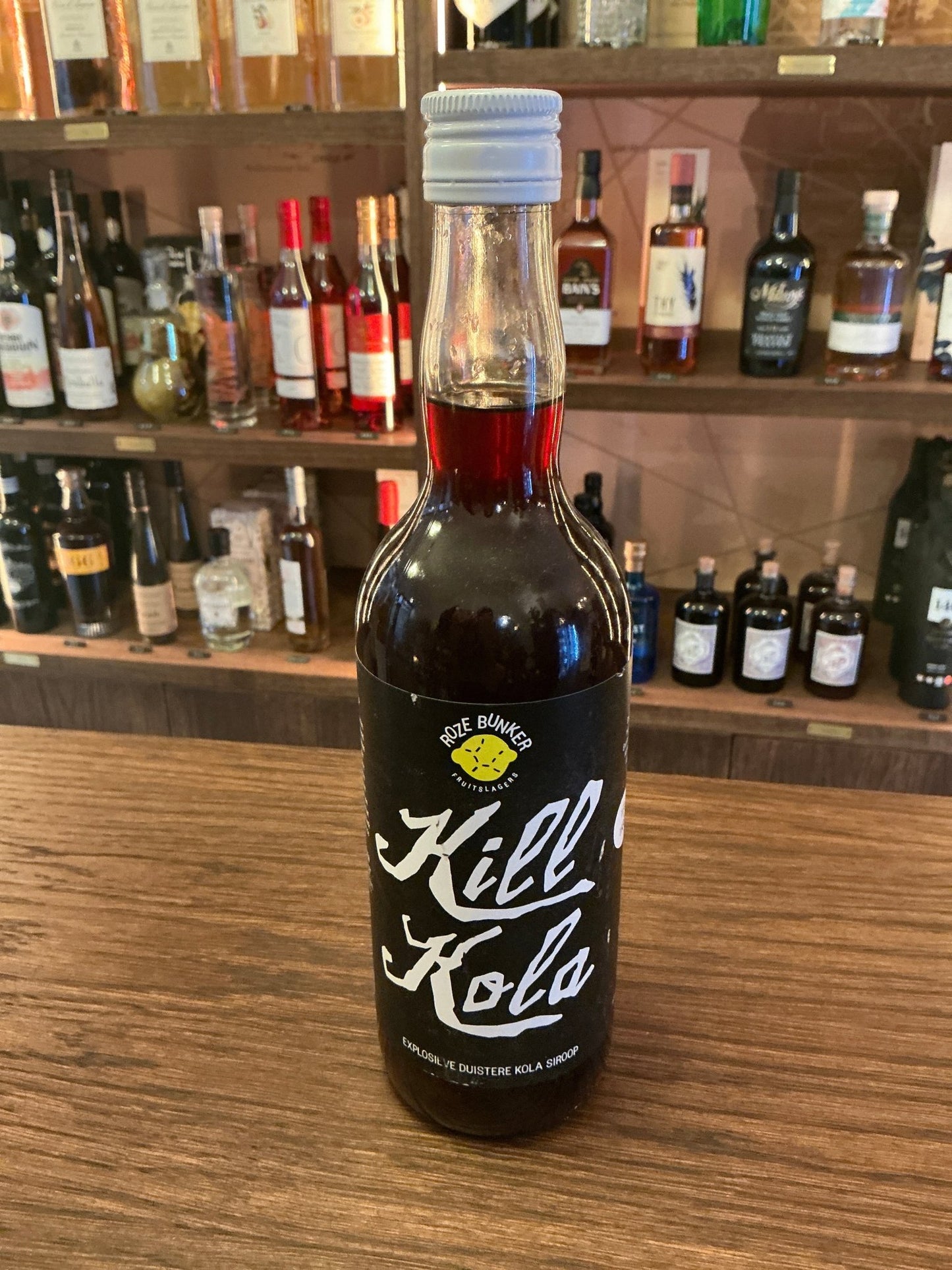 Kill Kola 690 ml dark spiced cordial bottle with white cap and bold white lettering on wooden bar counter with shelves of.