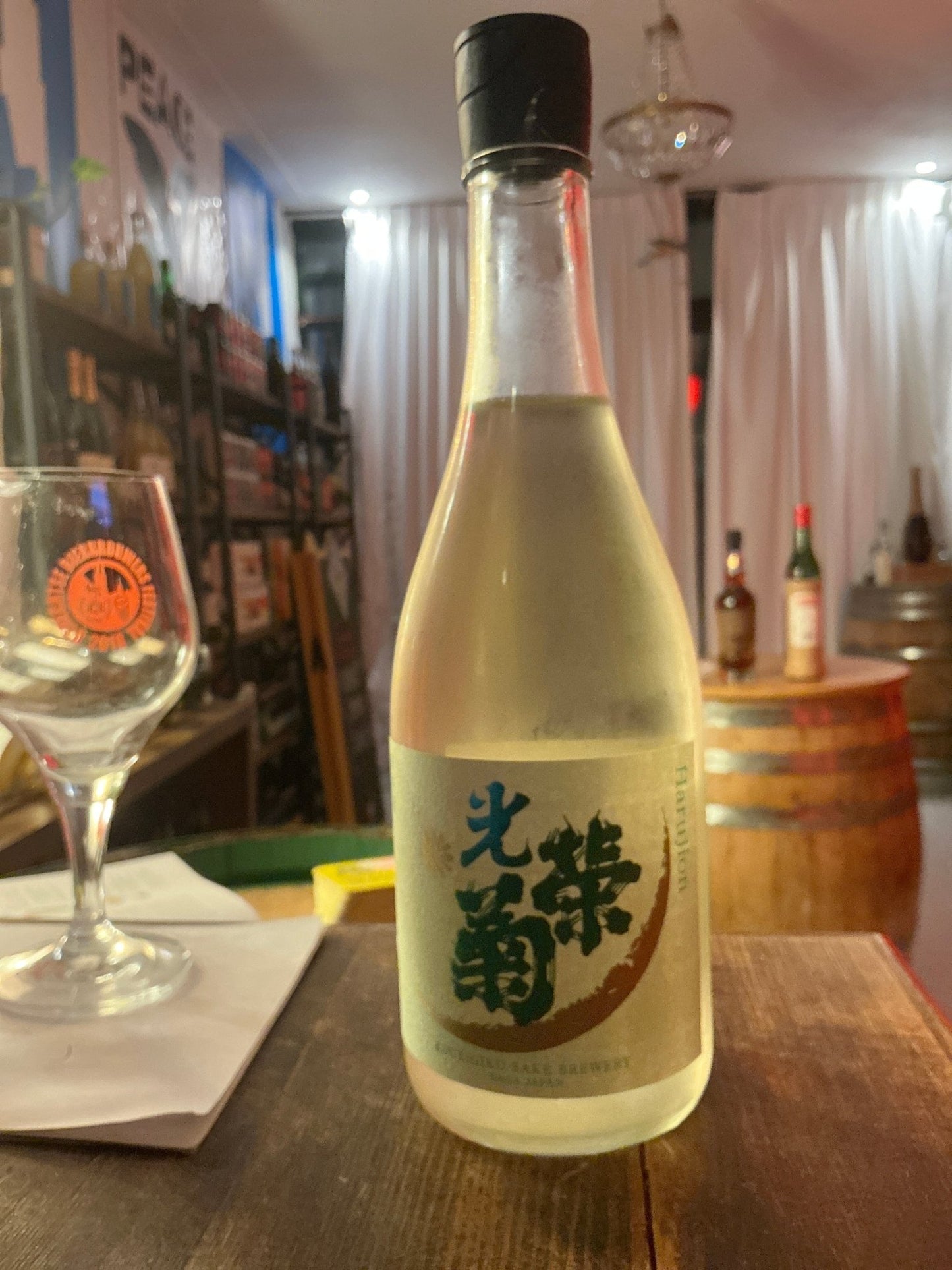 Clear glass bottle of Koueigiku Harujion sake with black cap and green label featuring Japanese characters, set on wooden.