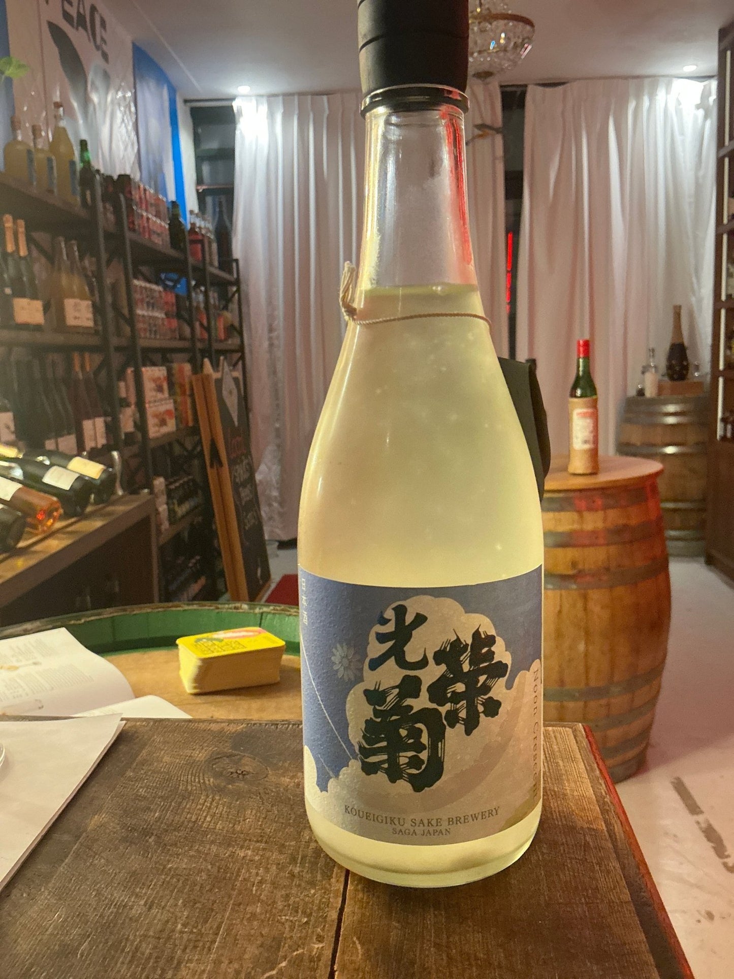 Koueigiku Noon Crescent premium sake bottle with pale yellow liquid and blue label featuring Japanese calligraphy on rustic.