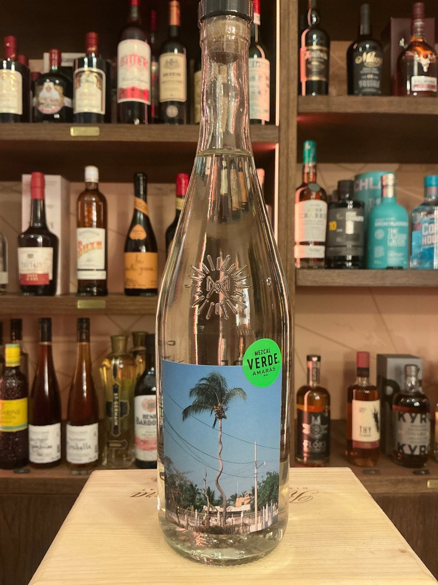 Clear glass bottle of Mezcal Verde Amarás with tall slender neck and tropical-themed label on wooden surface, surrounded by.