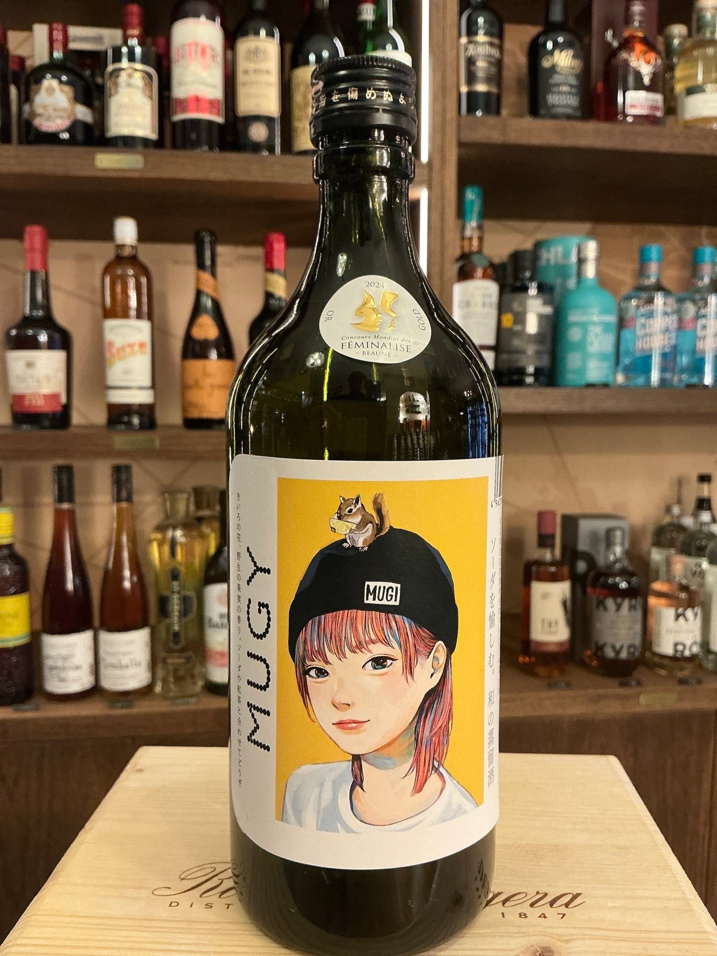Munemasa Mugi Shochu bottle with yellow label featuring illustration of a young woman in a black beanie, dark glass, artisan .