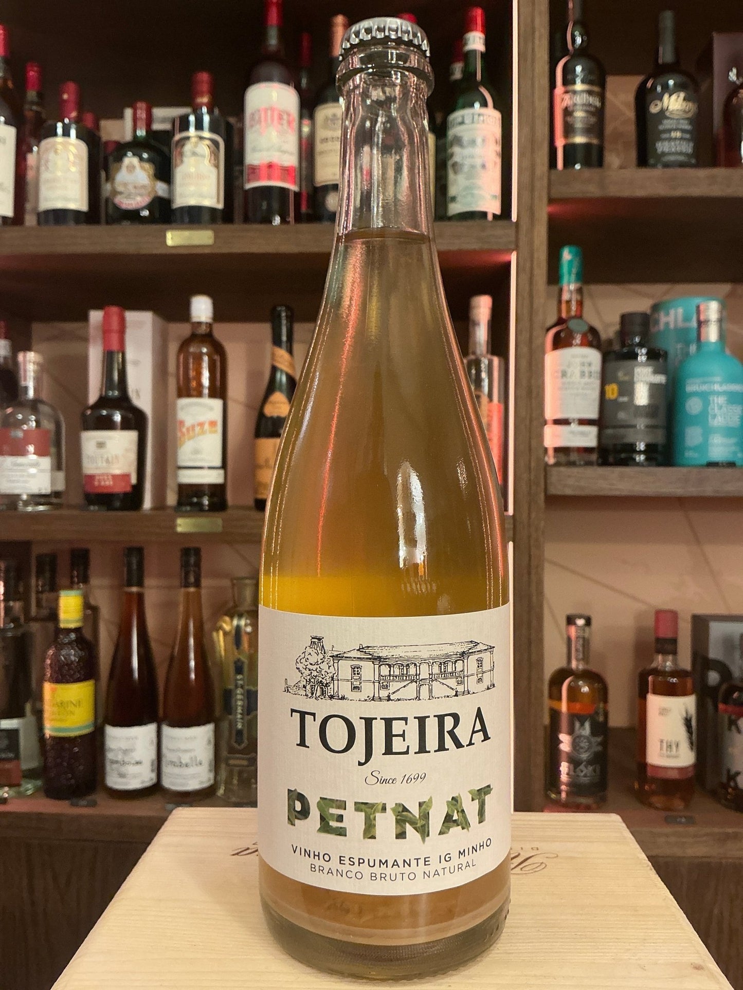 Casa da Tojeira Pét-Nat sparkling wine bottle with minimalist white label and black text on wooden surface in front of bar.