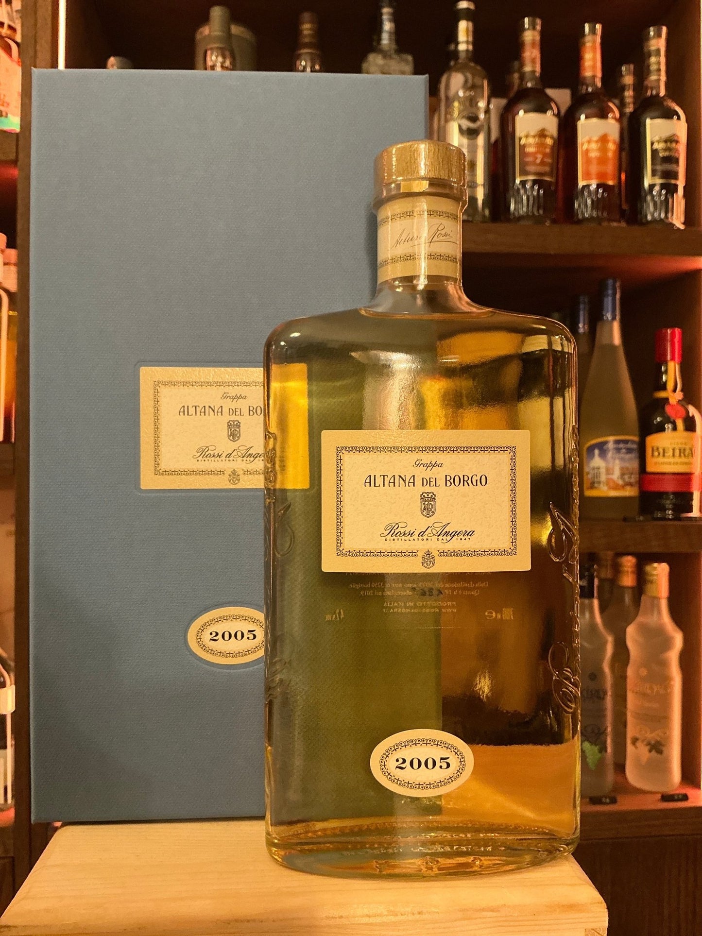 Rossi D’Angera Altana Del Borgo 2005 aged grappa bottle with beige label and wooden cap beside matching blue box on wooden.