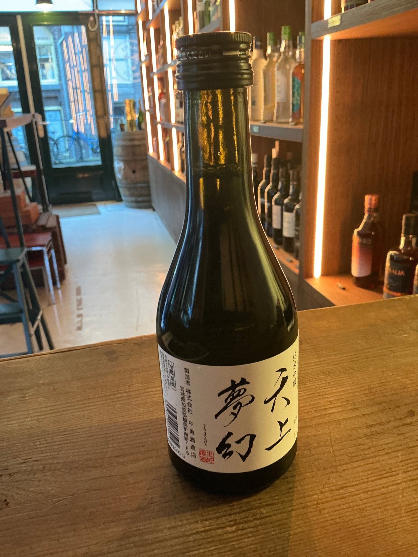 Tenjō Mugen Junmai Ginjo sake bottle with white label and black calligraphy on wooden bar counter, premium artisan distilled .