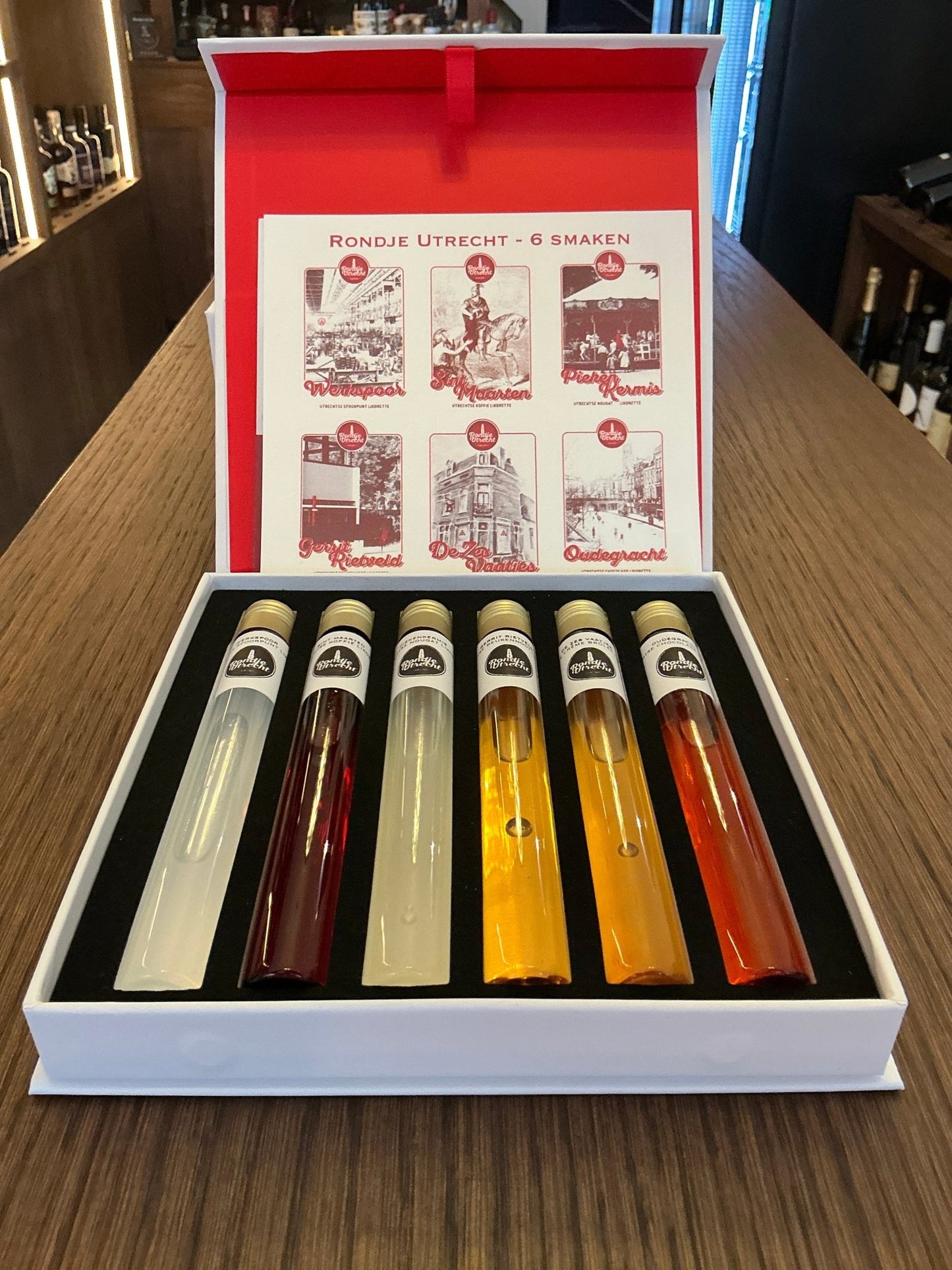 Open box with six tall, slender bottles of premium Dutch artisan liqueurs in clear glass, showcasing a variety of colors.