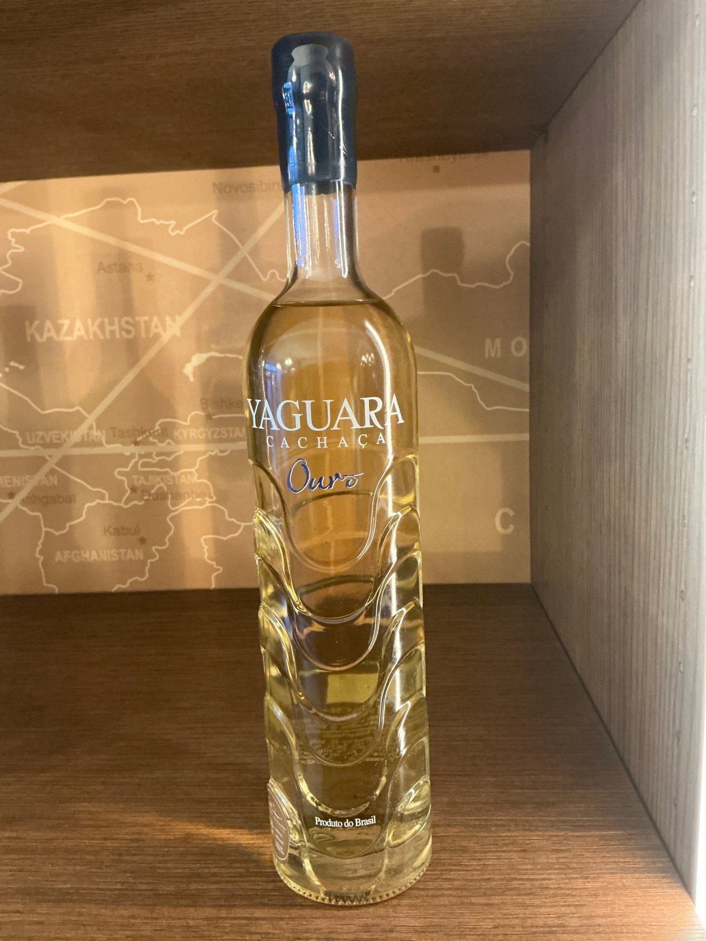 Tall, clear bottle of Yaguara Cachaca Ouro with gold spirit, blue foil seal, artisanal Dutch limited edition branding, on.