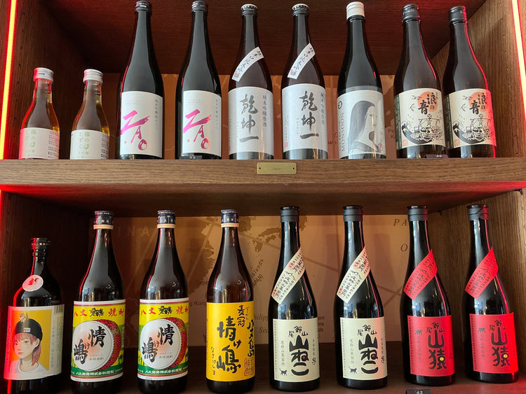 Japanese sake and shōchū bottles with illustrated labels arranged on two shelves