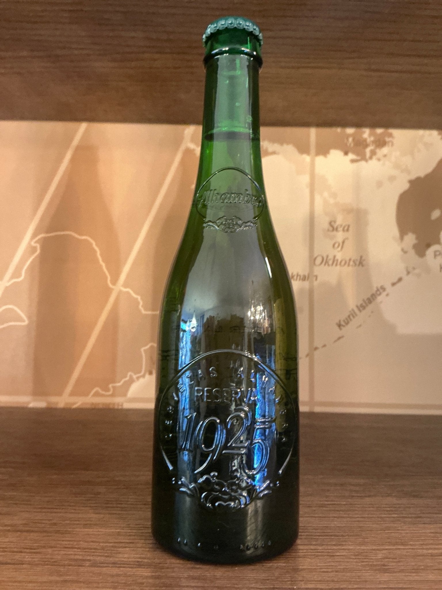 Tall green glass bottle with embossed decorative logo and text, capped with a matching green metal cap, set against a wooden