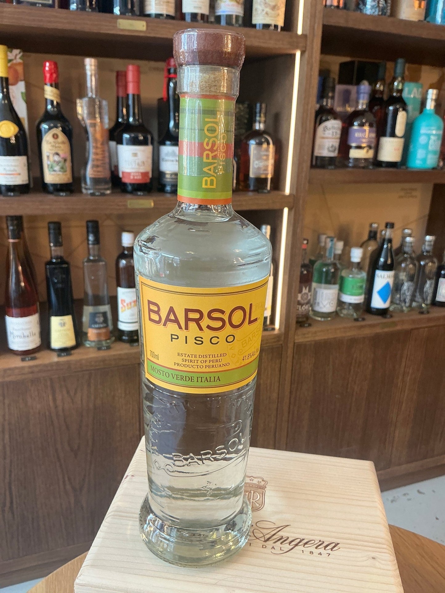 Barsol Mosto Verde Italia Pisco bottle with clear glass, yellow label, and wooden cap, displayed on wooden surface with.