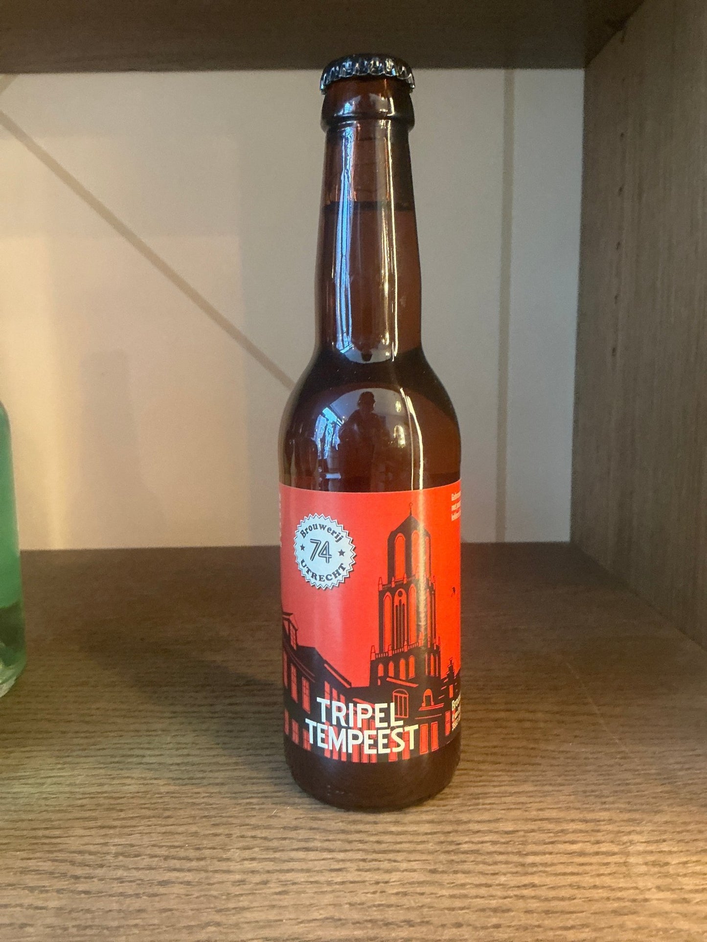 Brown glass bottle of Brouwerij 74 Tripel Tempeest Belgian Tripel with a vibrant orange label, featuring Dutch cityscape.