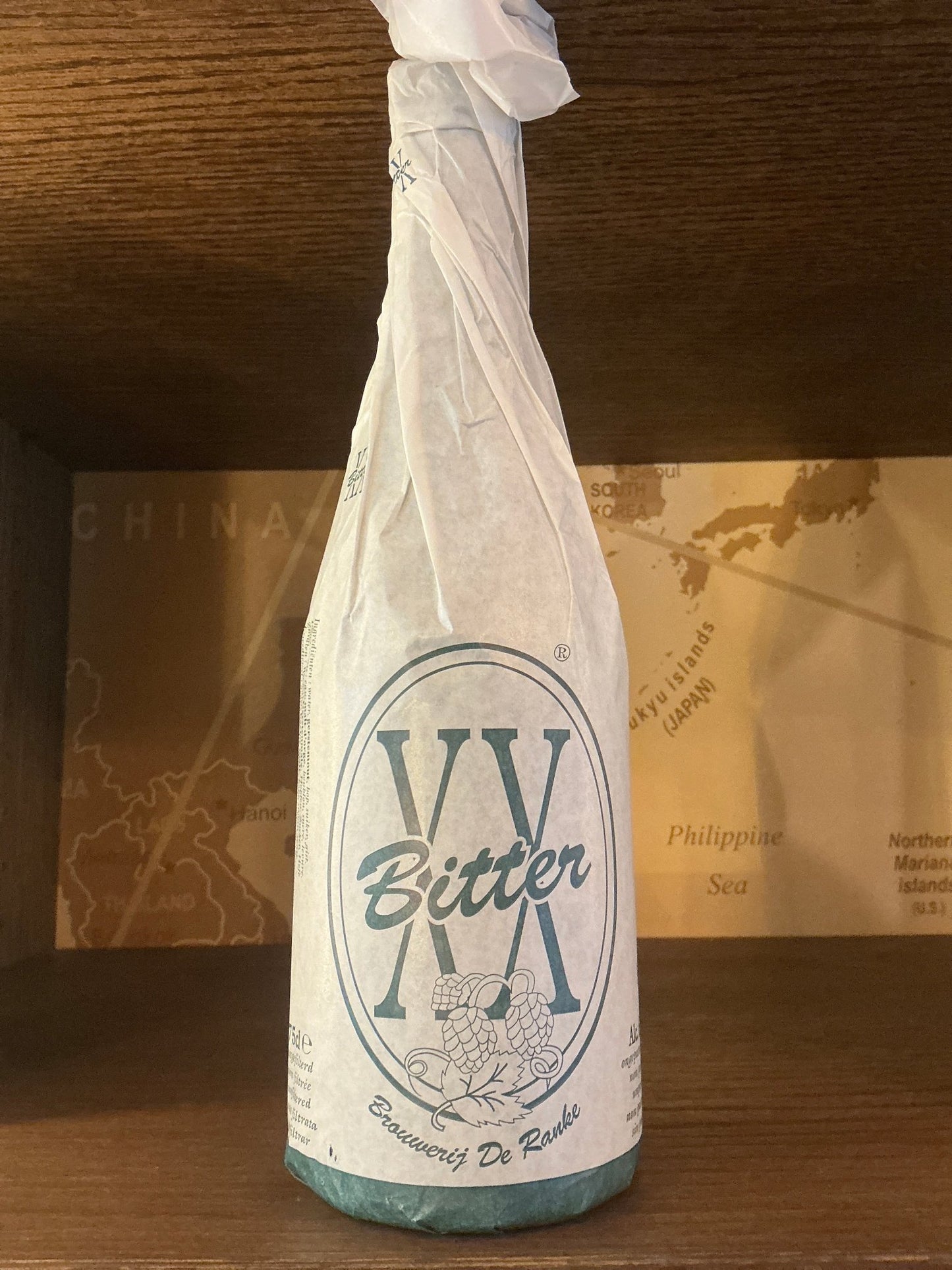 Brouwerij De Ranke XX Bitter craft beer bottle wrapped in white paper with minimalist blue logo and text on wood surface.