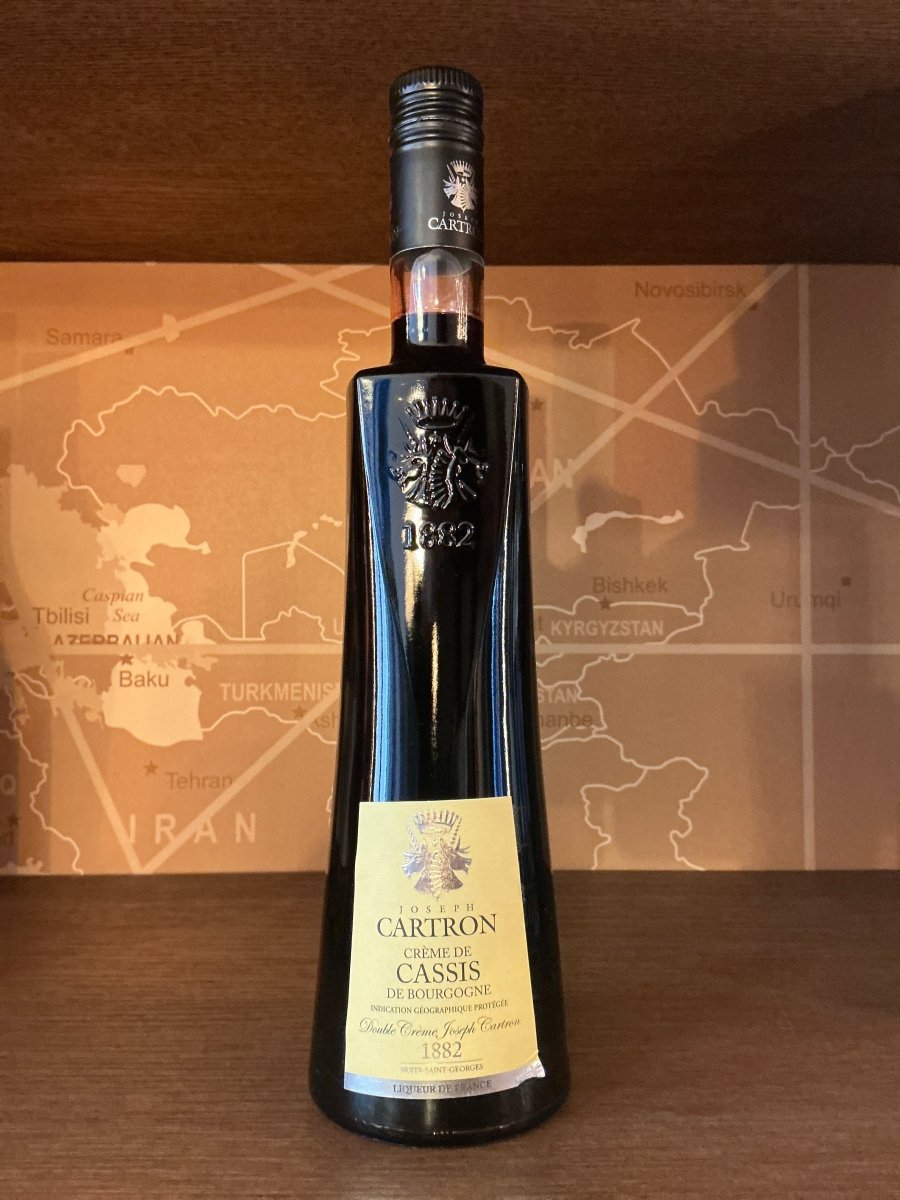 Tall dark glass bottle of Carton Crème de Cassis liqueur with botanical and artisan styling on label, displayed against.