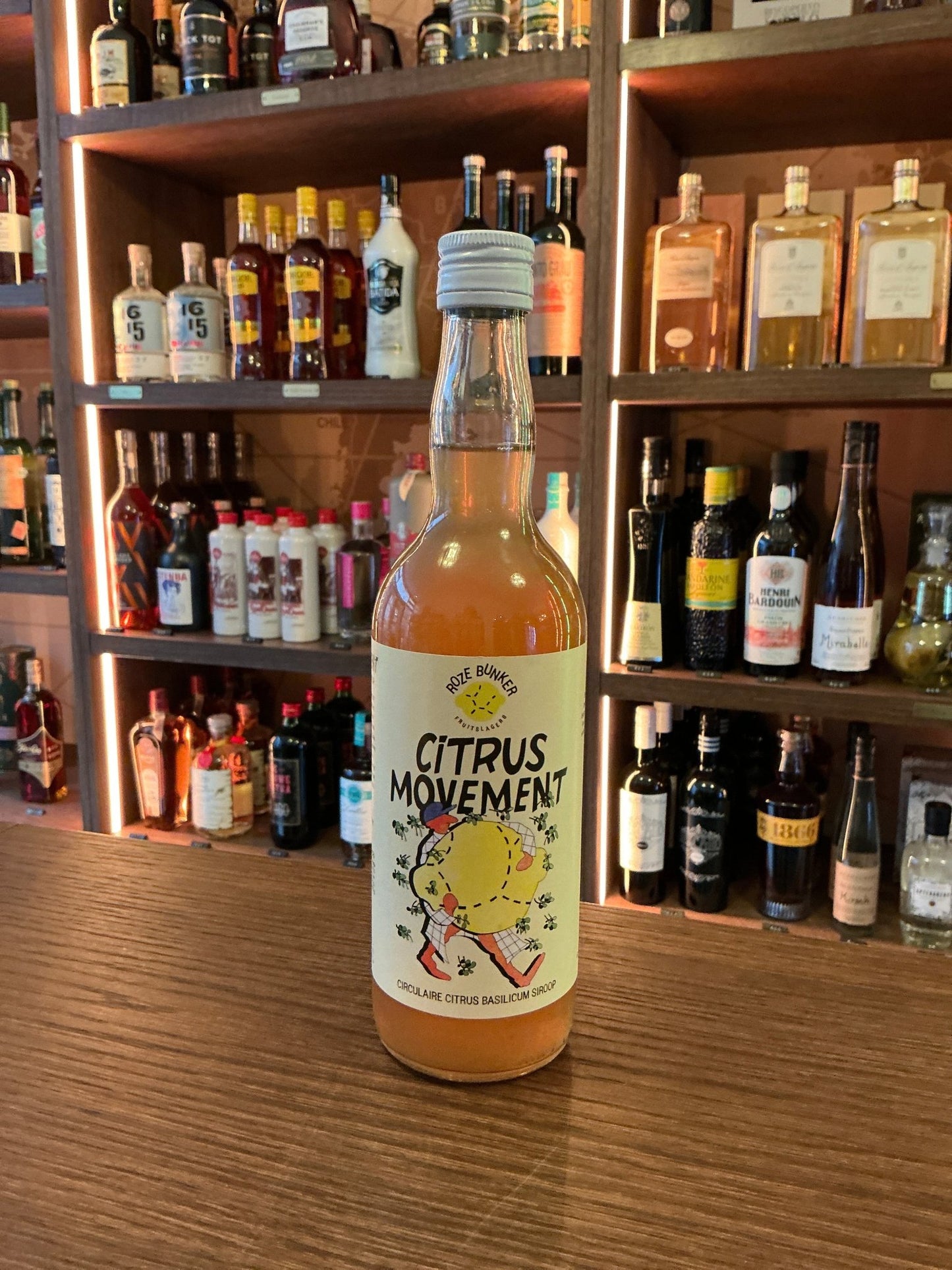 Citrus Movement Citrus & Basil Syrup in a clear glass bottle with white cap, displayed on a wooden bar counter with premium.