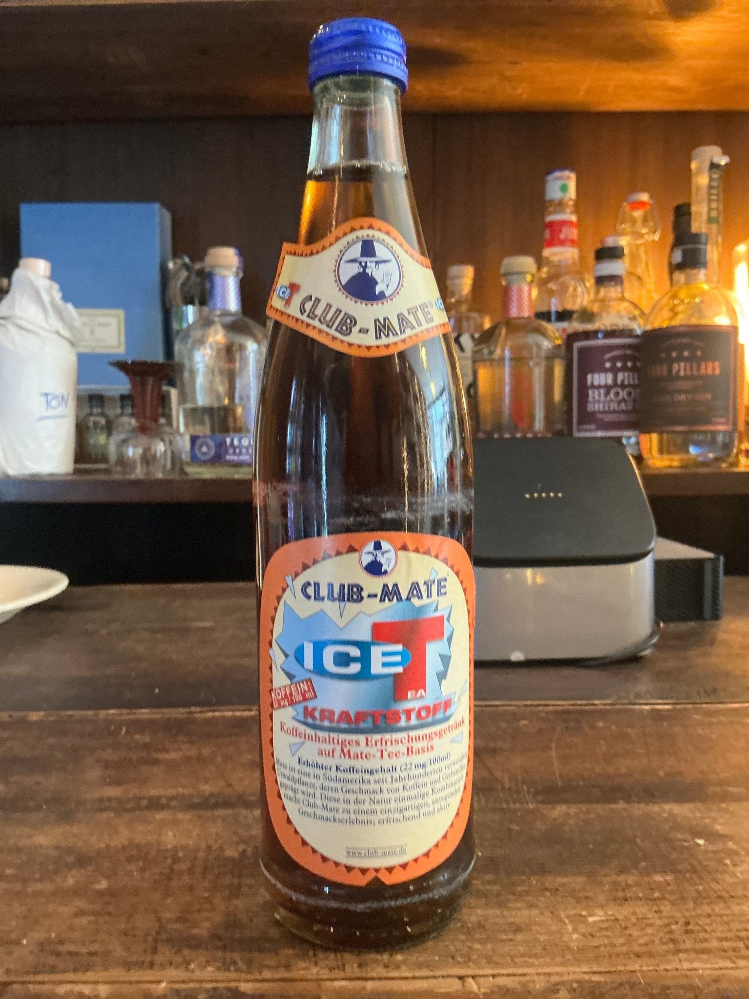 Club Mate Ice Tea Kraftstoff bottle with blue cap on wooden bar counter, premium artisan botanical soda beside assorted.