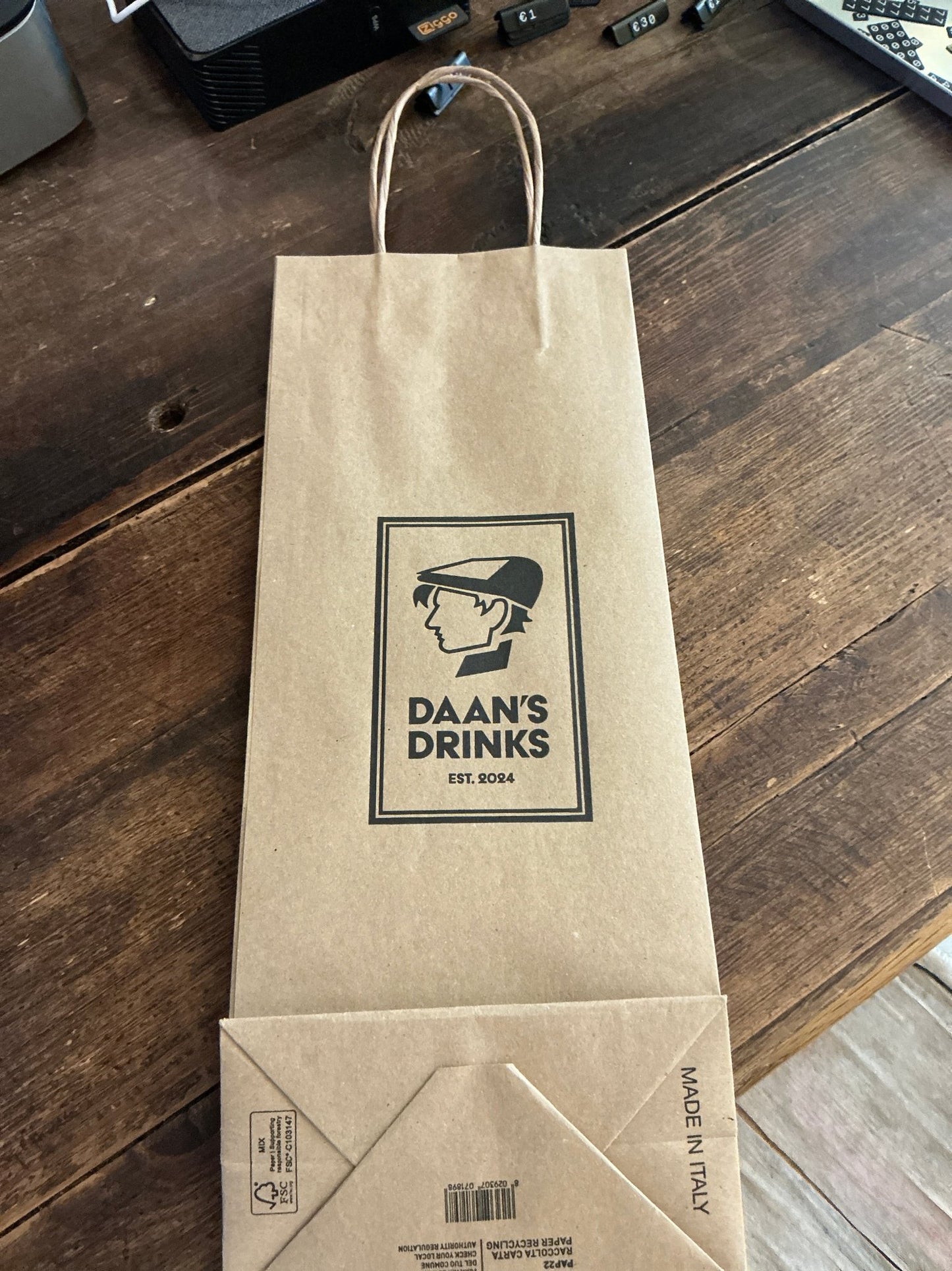 Natural brown kraft paper bag with twisted handles, featuring black logo of Daan's Drinks and vintage-styled illustration,.