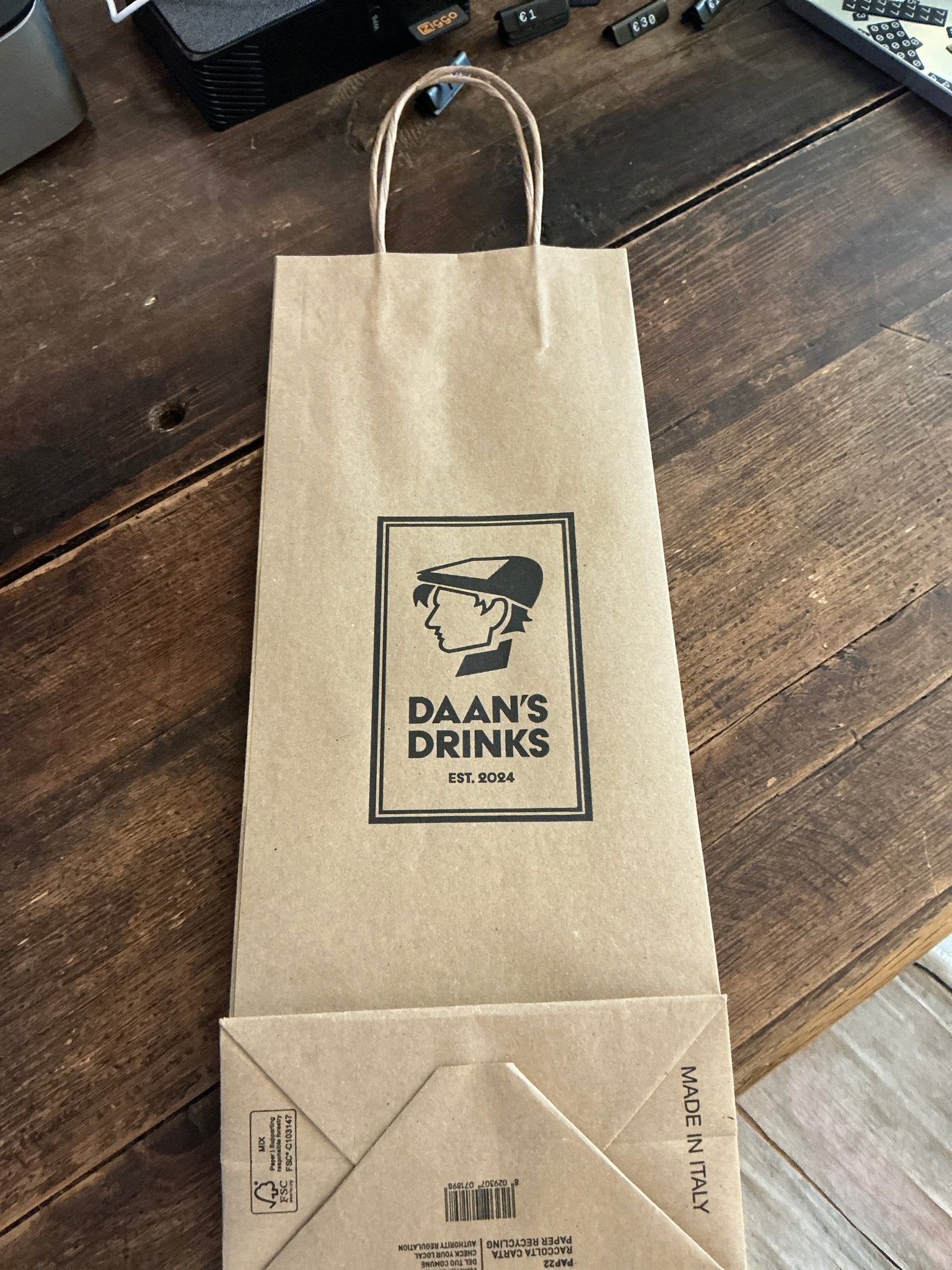 Natural brown kraft paper bag with twisted handles, featuring black logo of Daan's Drinks and vintage-styled illustration,.