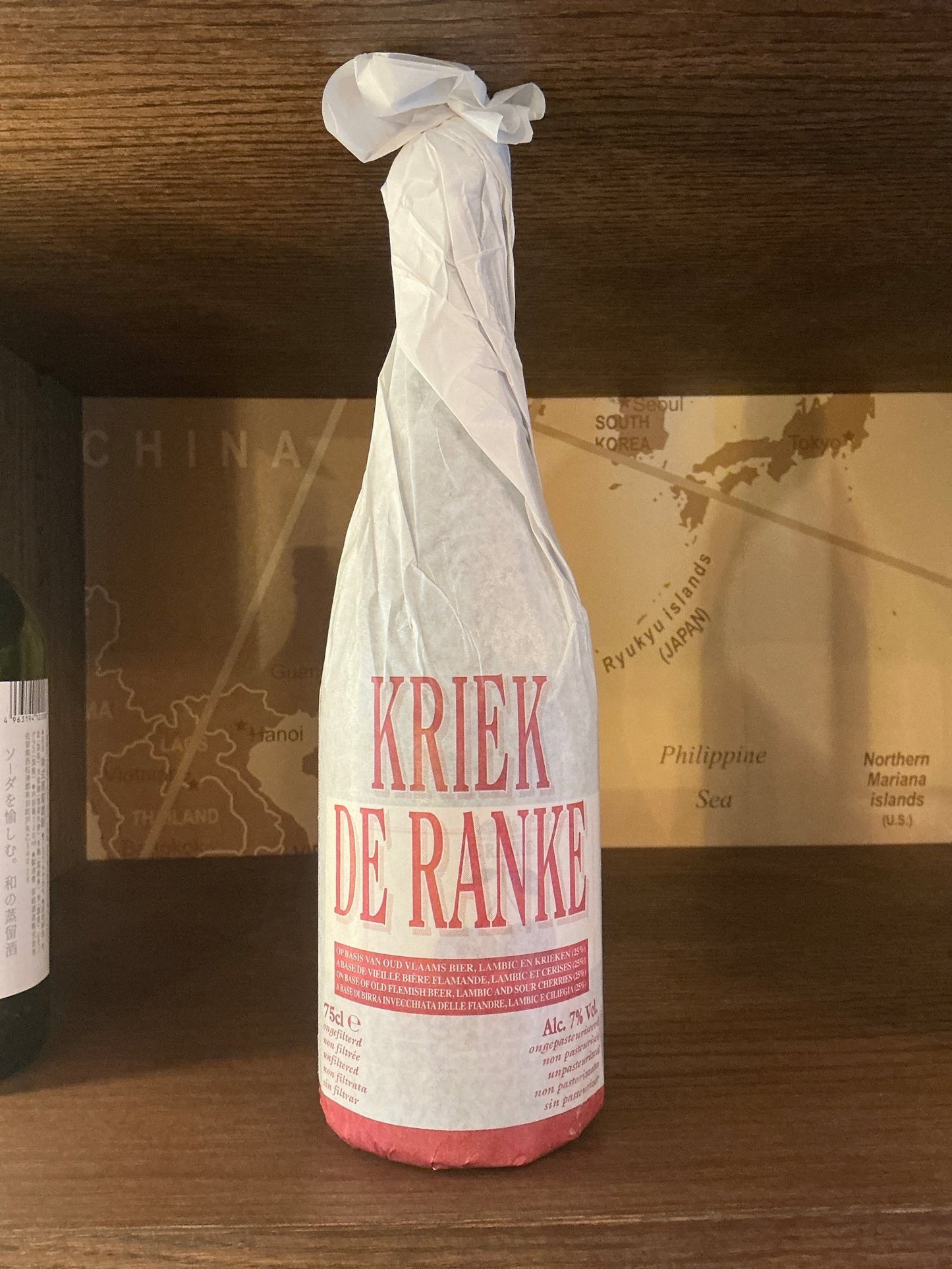 Tall bottle wrapped in white paper with red text reading Kriek De Ranke, artisan small batch specialty spirits on wooden.