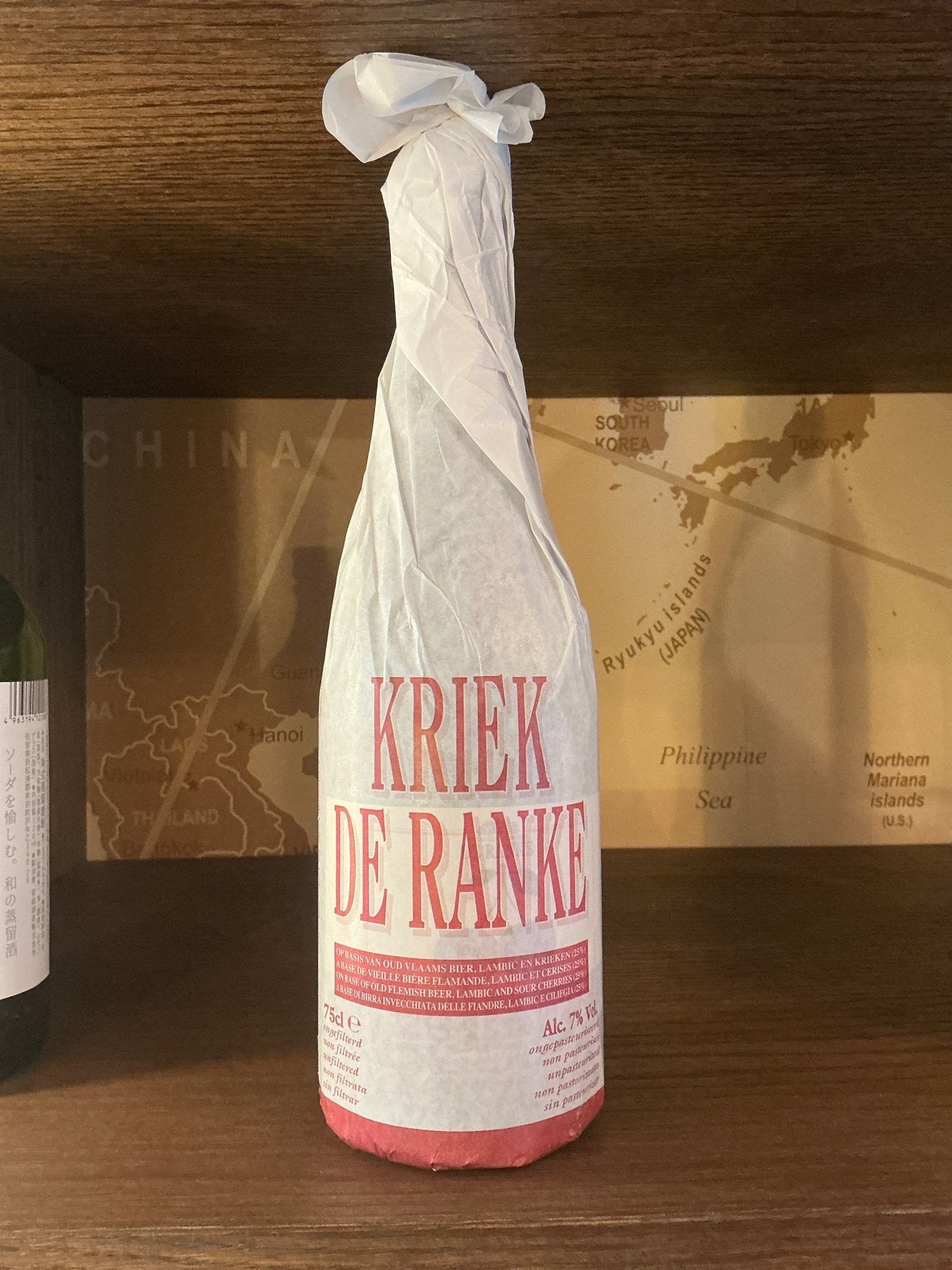Tall bottle wrapped in white paper with red text reading Kriek De Ranke, artisan small batch specialty spirits on wooden.