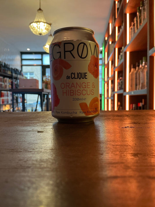Degrow Lab De Clique Orange Hibiscus kombucha can with botanical illustration on wooden table in artisanal beverage shop.