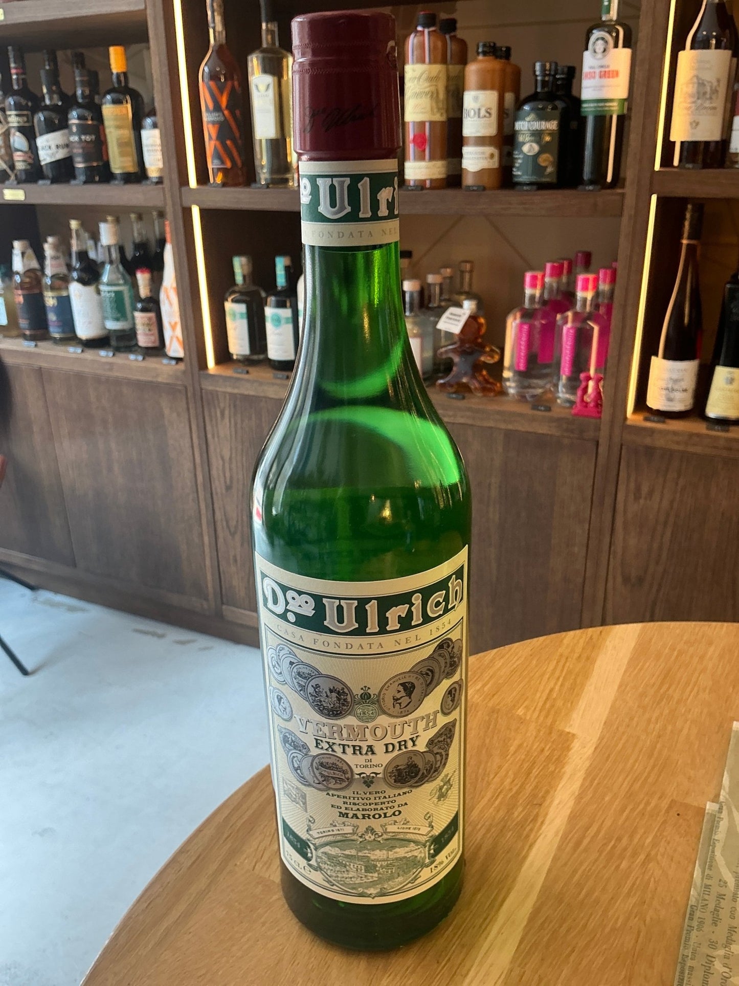 Dr Ulrich Vermouth Extra Dry green glass bottle with vintage botanical label on wooden table in rustic bar setting.