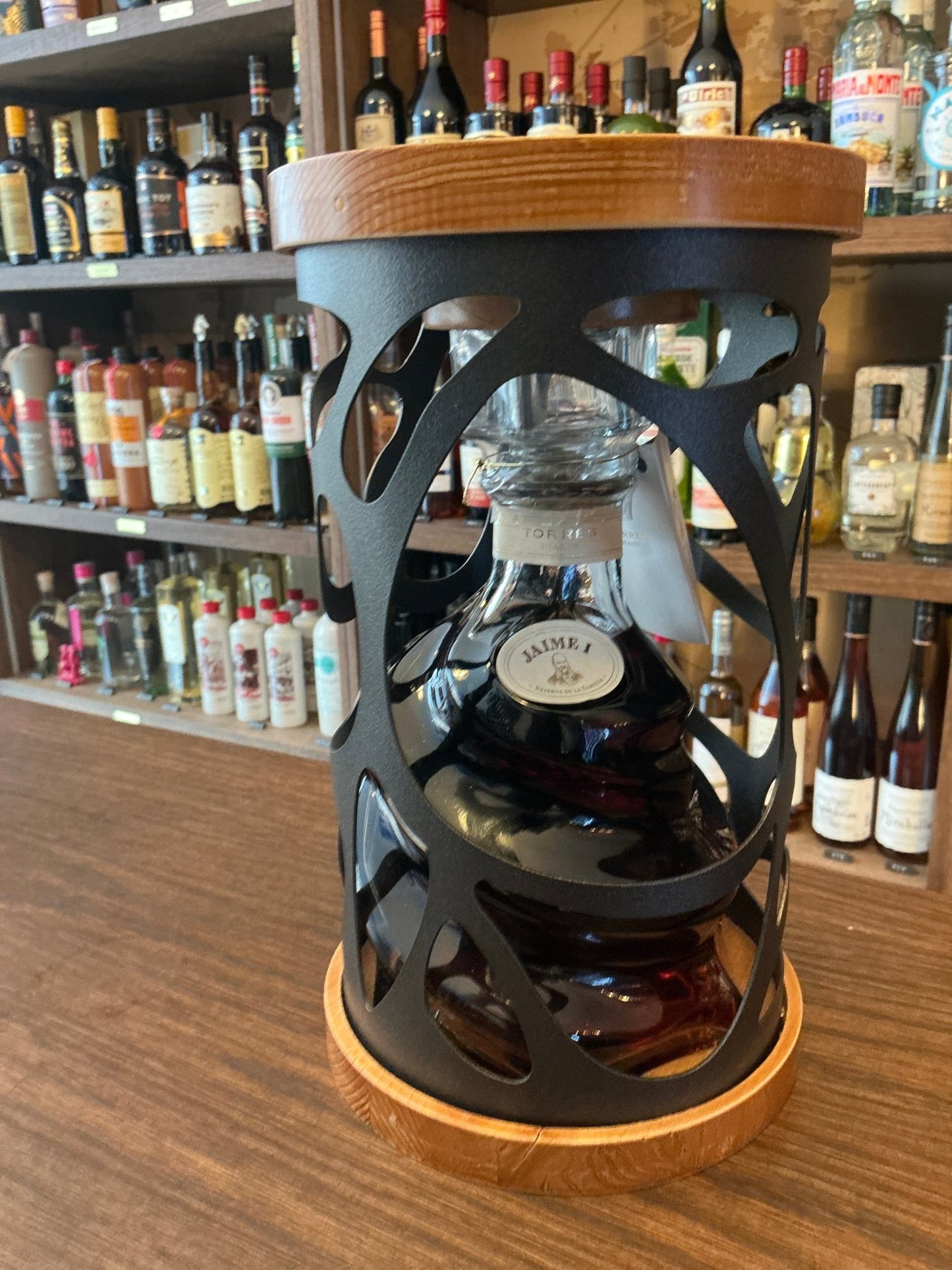 Premium Jaime I Brandy in an artisan glass bottle with wooden top and base, displayed on a wooden counter with shelves of.