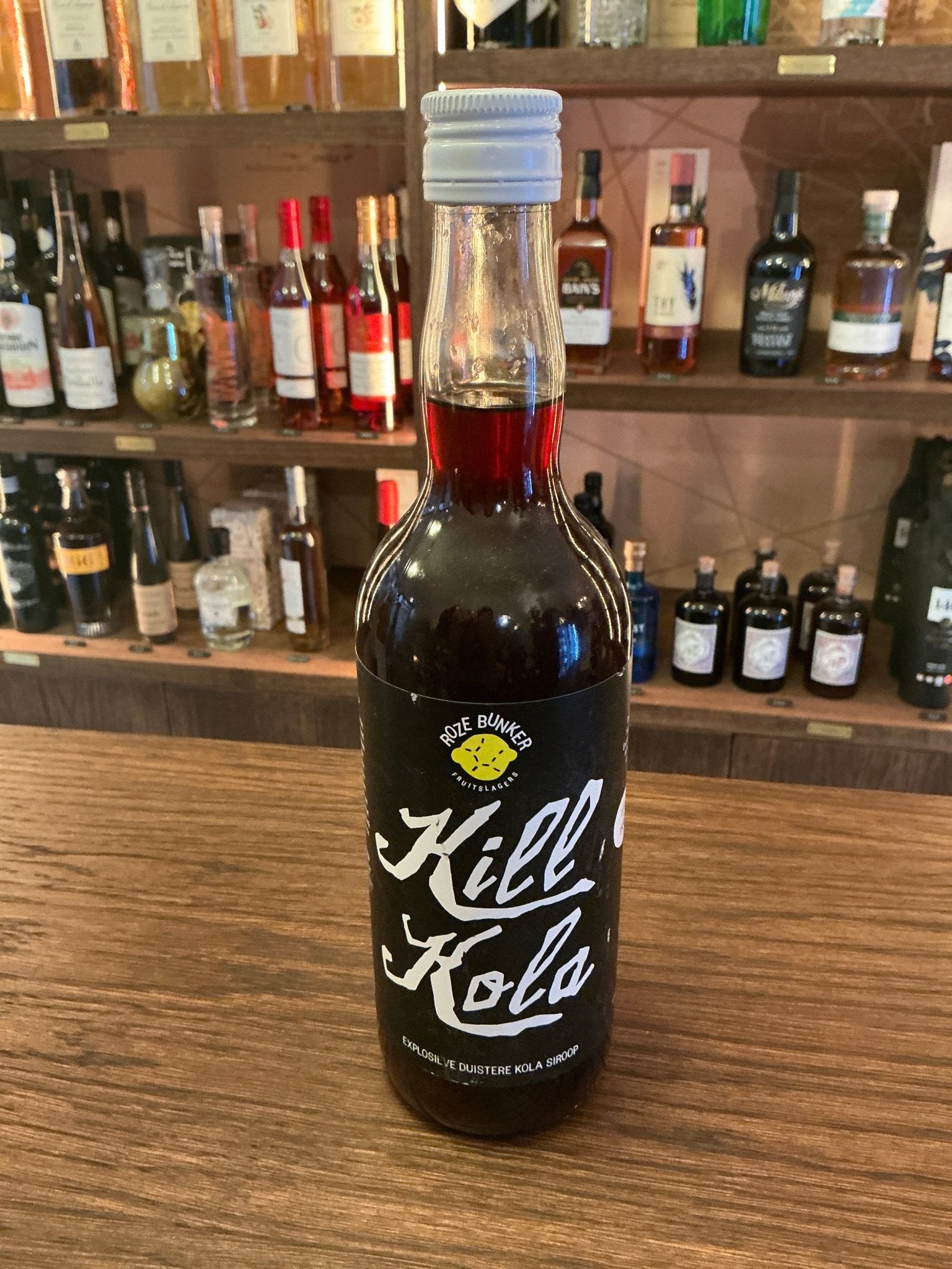 Kill Kola 690 ml dark spiced cordial bottle with white cap and bold white lettering on wooden bar counter with shelves of.