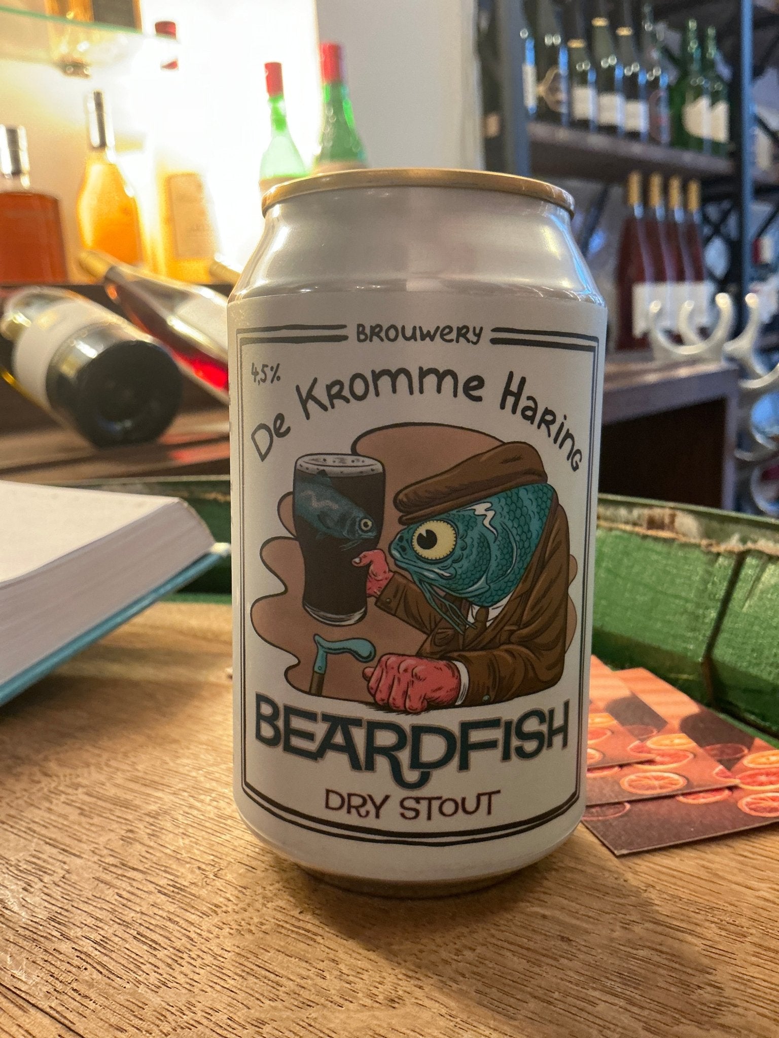 White can of Kromme Haring Beardfish Dry Stout craft beer with colorful cartoon fish logo on wooden table in a bar setting.