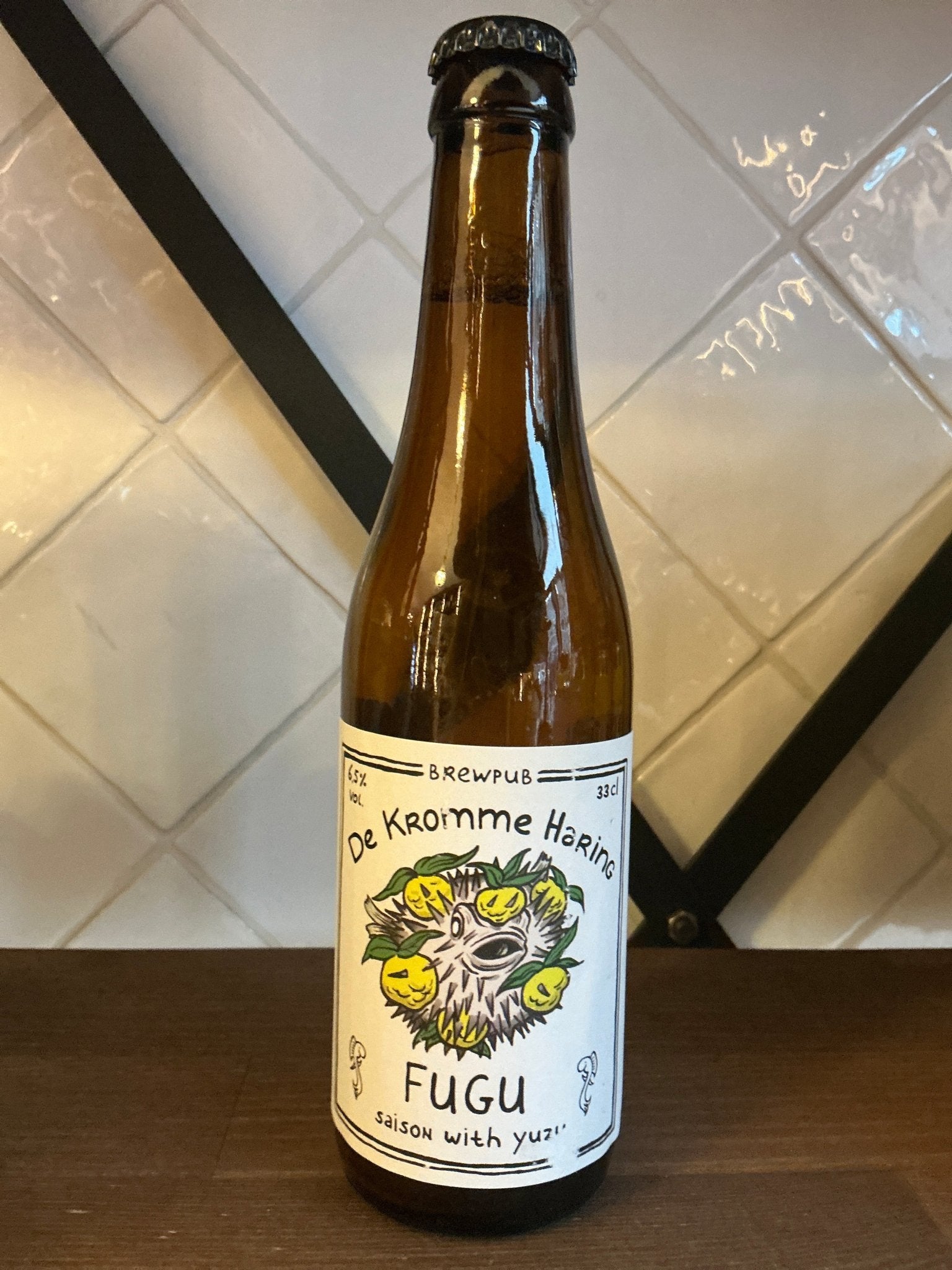 Kromme Haring Fugu Saison craft beer bottle with botanical yuzu label on dark glass against tiled background.