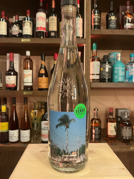 Clear glass bottle of Mezcal Verde Amarás with tall slender neck and tropical-themed label on wooden surface, surrounded by.