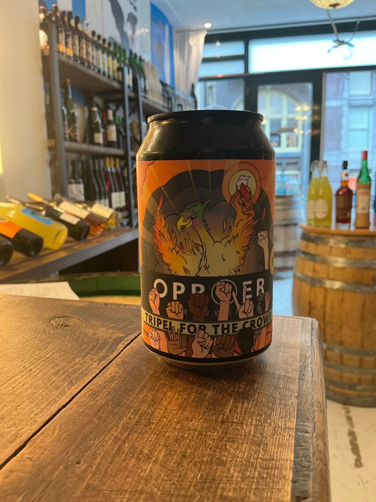 Oproer Tripel For The Crowd Belgian ale craft beer can with vibrant orange and dark botanical-themed label on wooden table.