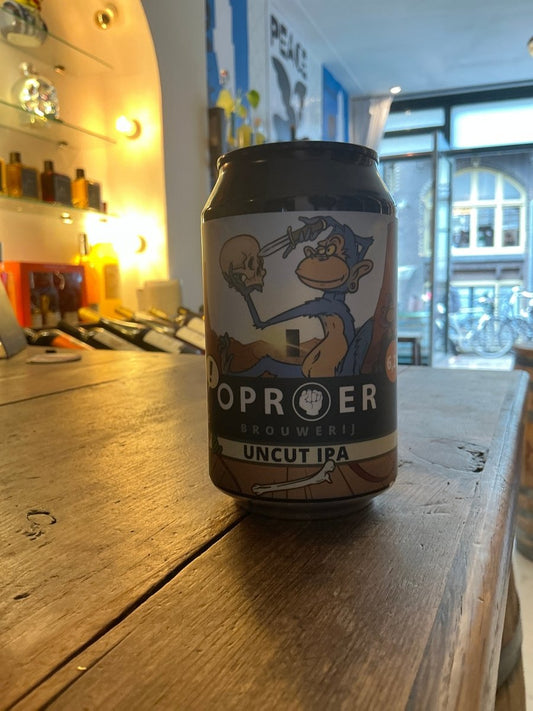 Oproer Uncut IPA craft beer can with cartoon fox holding a cup, black and white label on wooden table in cozy bar setting.