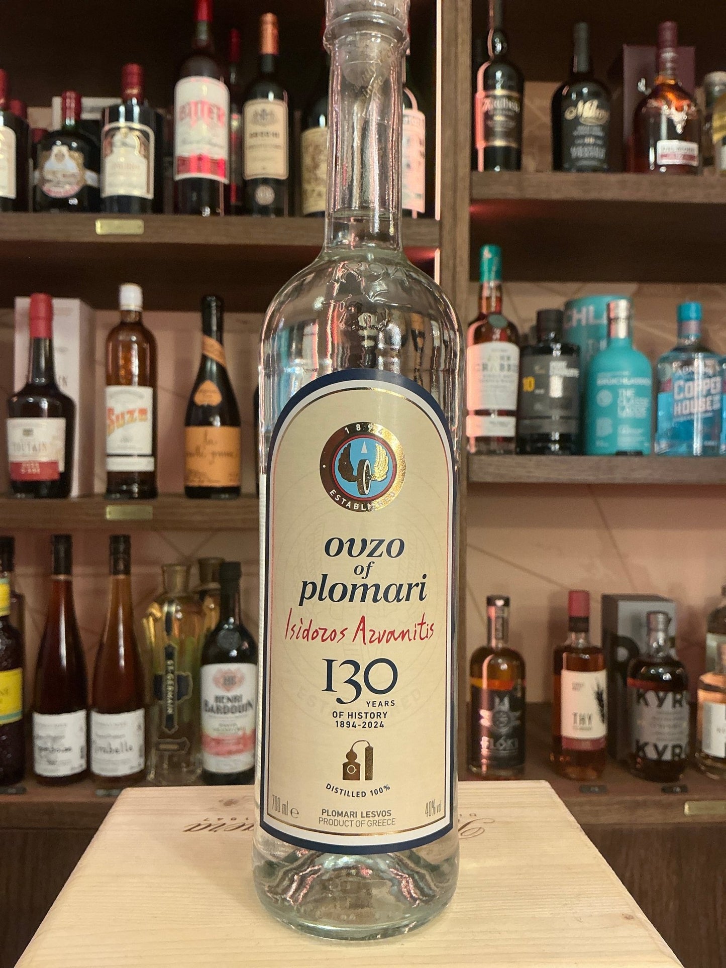 Clear glass bottle of Ouzo Plomari Greek aniseed-flavored liqueur with cream label, black and gold detailing, standing on.