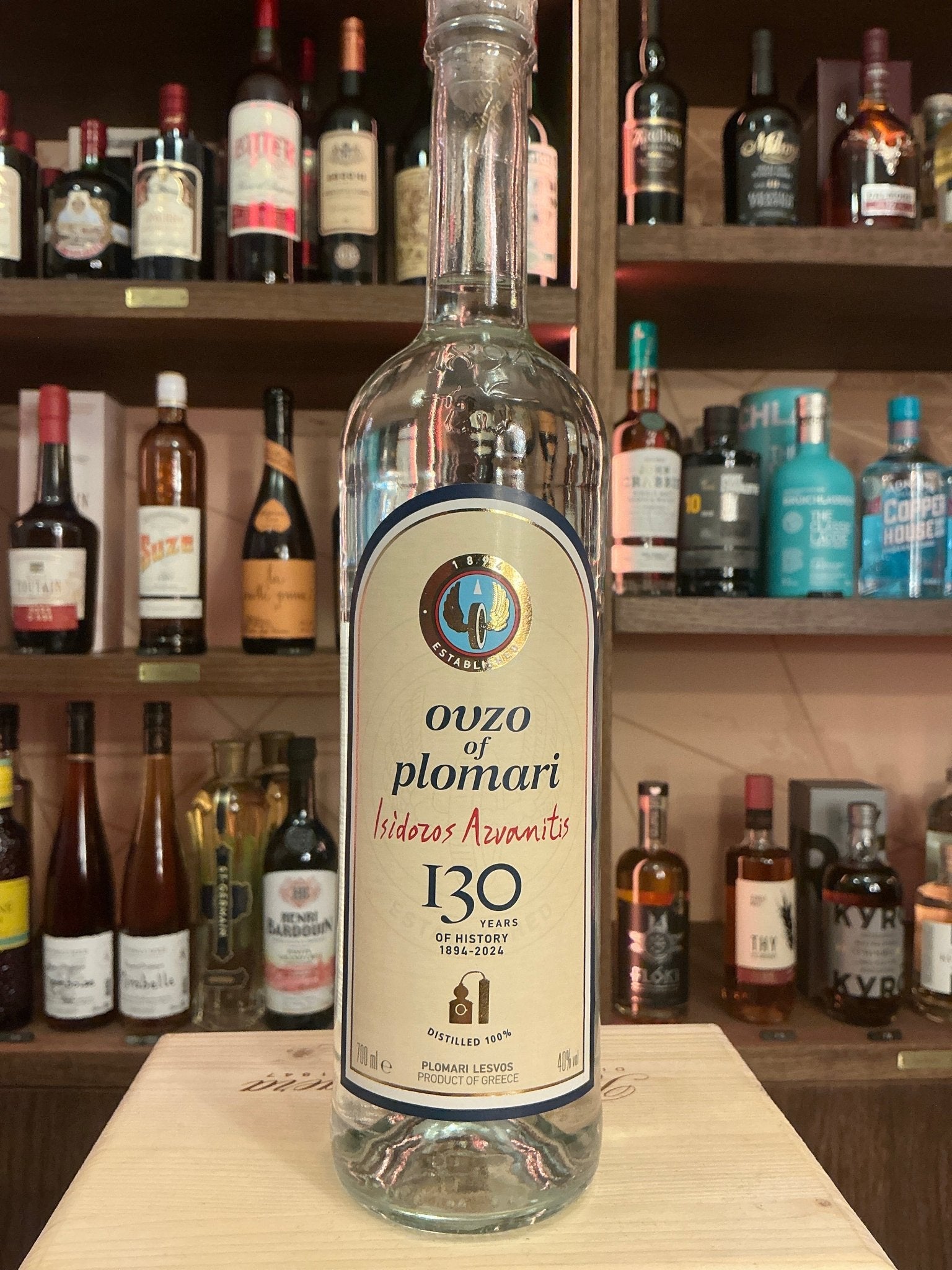 Clear glass bottle of Ouzo Plomari Greek aniseed-flavored liqueur with cream label, black and gold detailing, standing on.