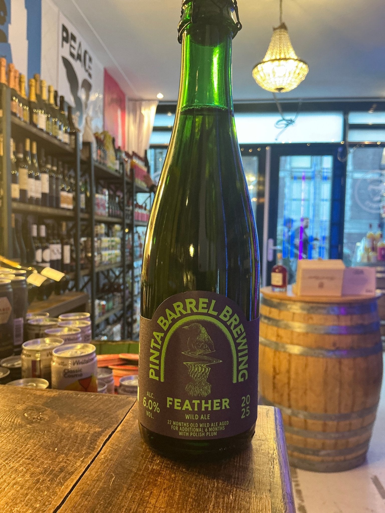 Green glass bottle of Pinta Barrel Brewing Feather wild ale with purple and gold label on wooden table in artisanal shop.
