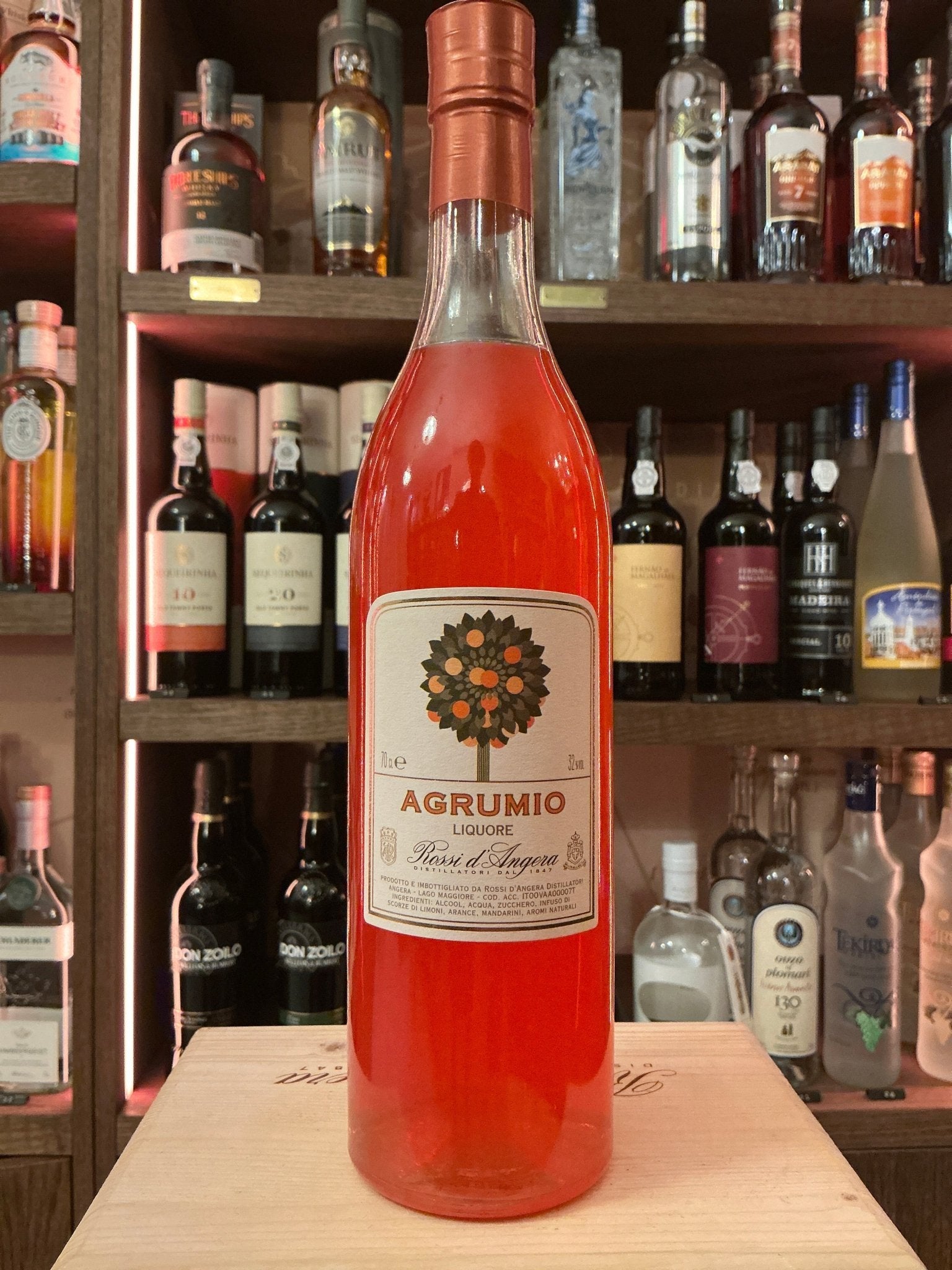 Tall clear glass bottle of Rossi D'Angera Agrumio citrus liqueur with copper cap and detailed botanical label on wooden.