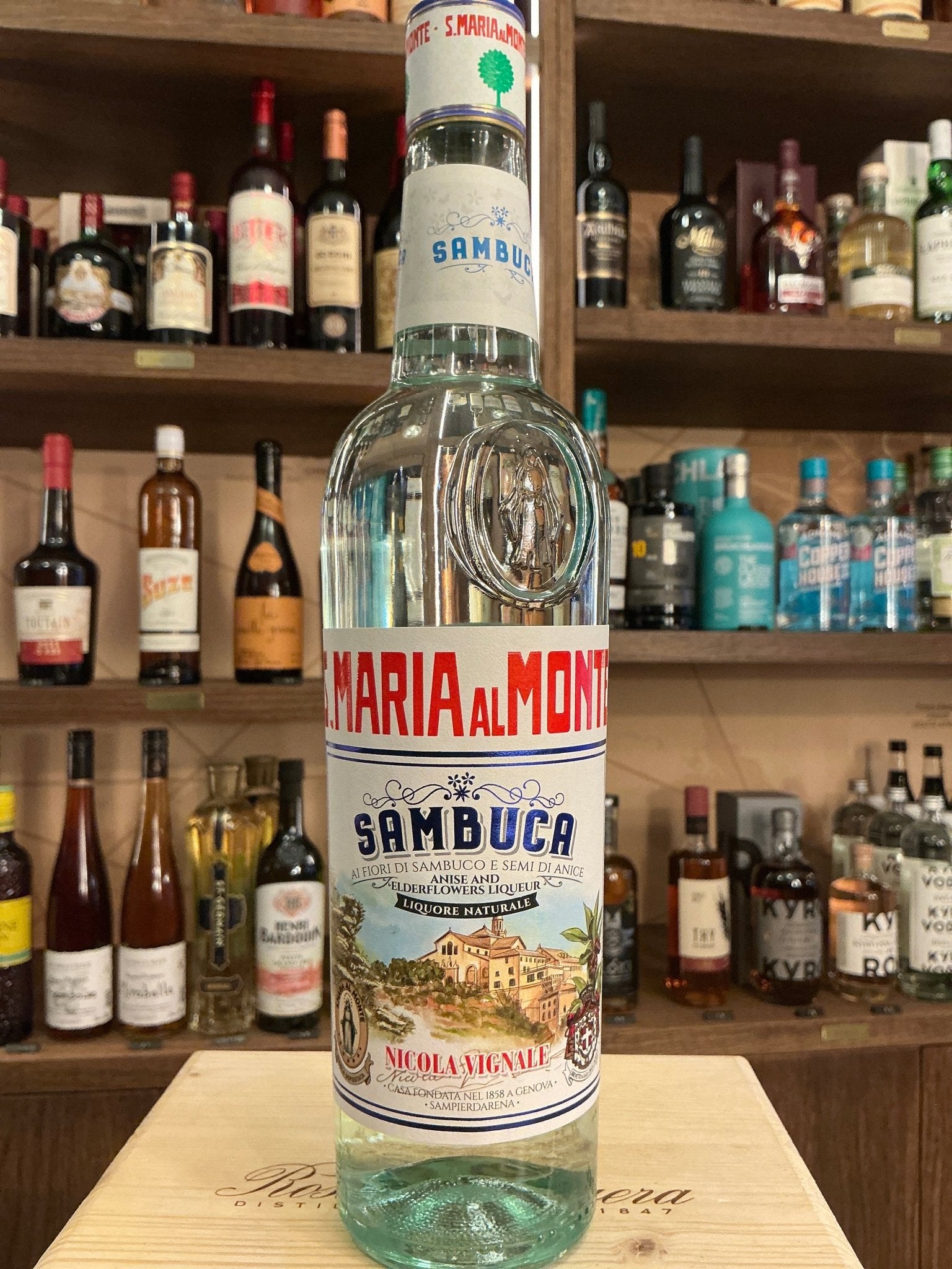 Clear Santa Maria Al Monte Sambuca bottle with blue and red label, artisan Italian liqueur, botanical and spiced flavors,.