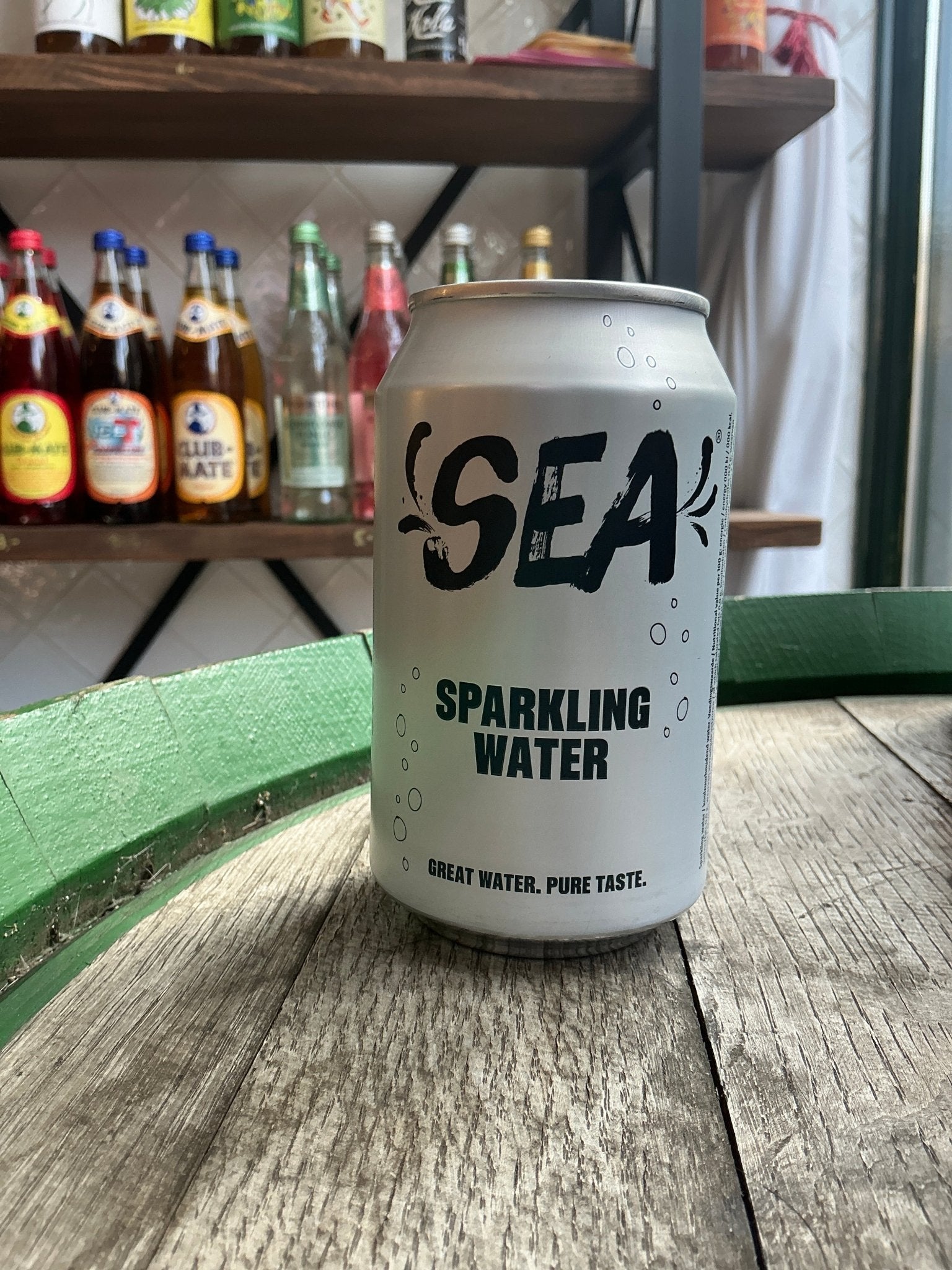 Sea sparkling water can with minimalist black text on white, set on rustic wood with artisan spirits in background.