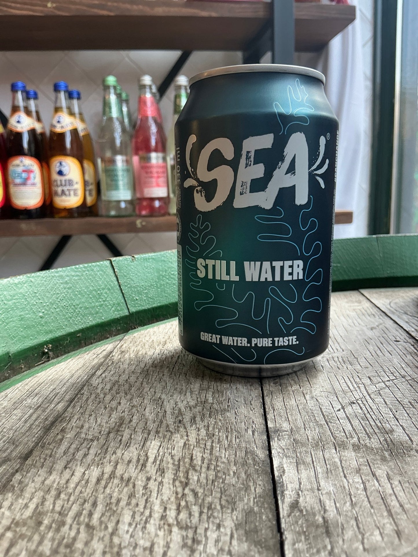Dark green can of Sea Still Water sparkling water with light blue and white branding, placed on weathered wooden surface.