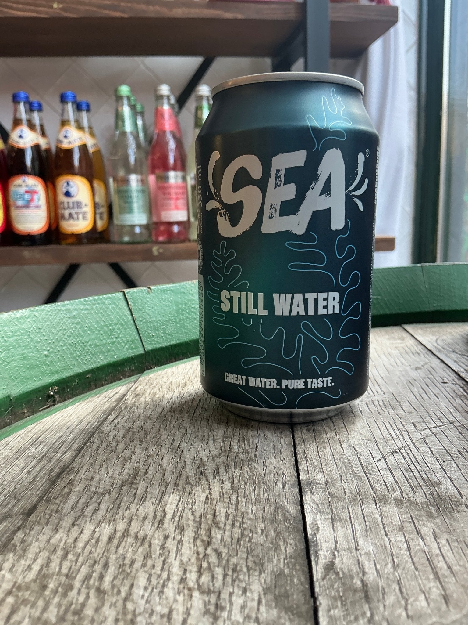 Dark green can of Sea Still Water sparkling water with light blue and white branding, placed on weathered wooden surface.