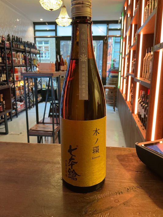 Tall amber glass bottle of Shichihonyari Junmai-shu Kinowa sake with yellow label featuring bold Japanese calligraphy, set.