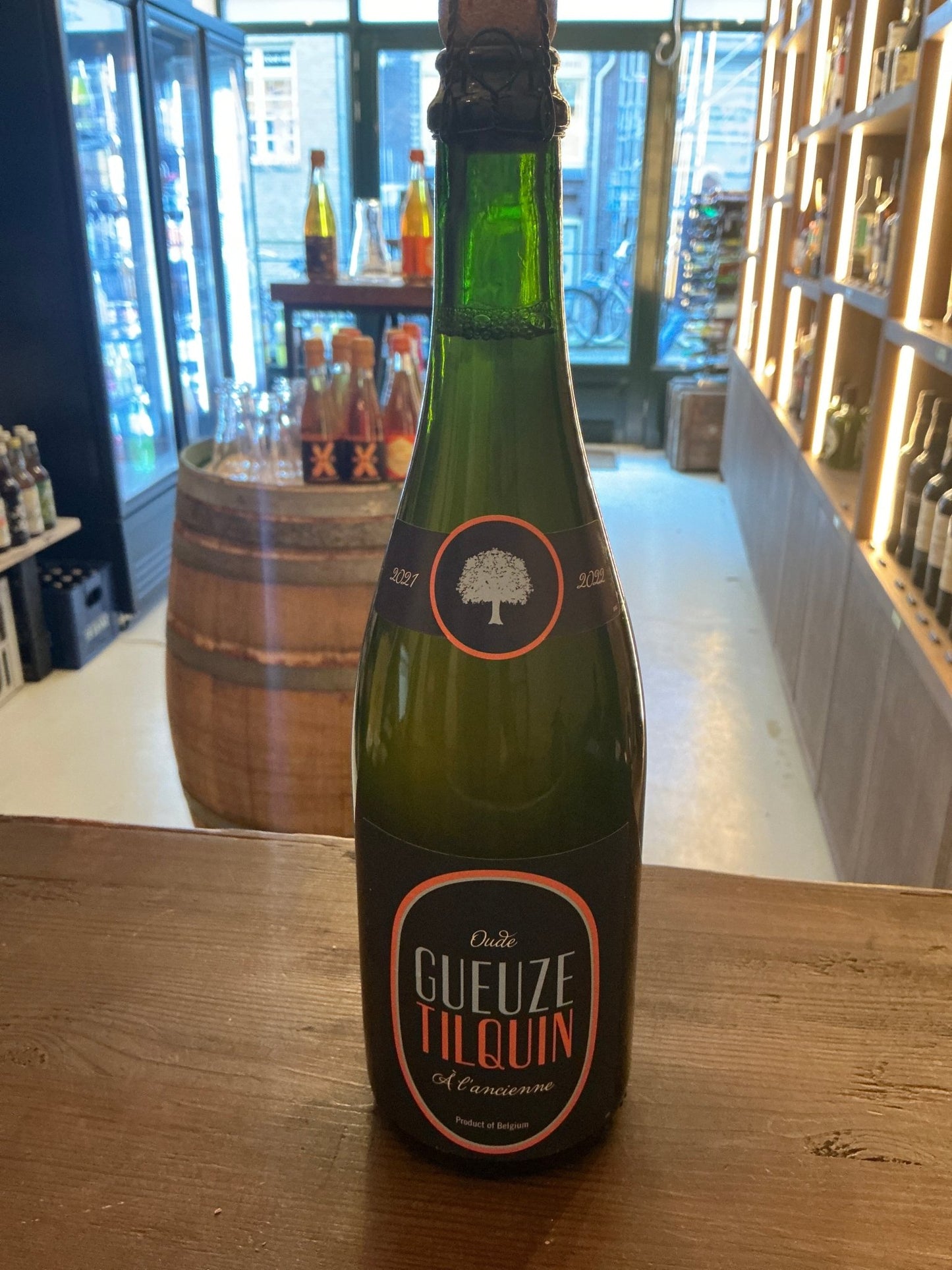 Green glass bottle of Tilquin Oude Gueuze craft beer with black and orange label, displayed in a specialty spirits shop with .